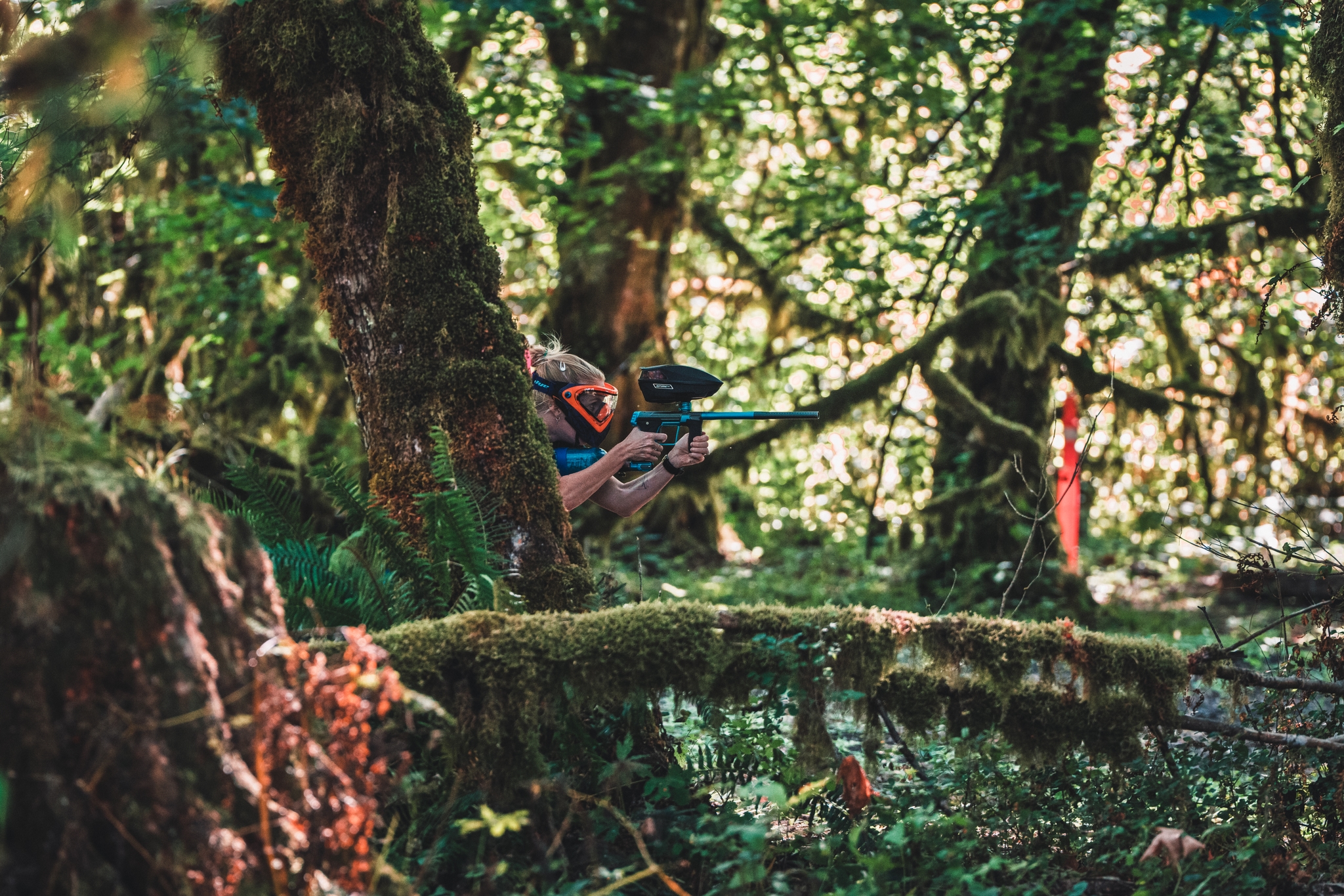 A person wearing a mask and holding a paintball gun is crouched in a dense, green forest setting