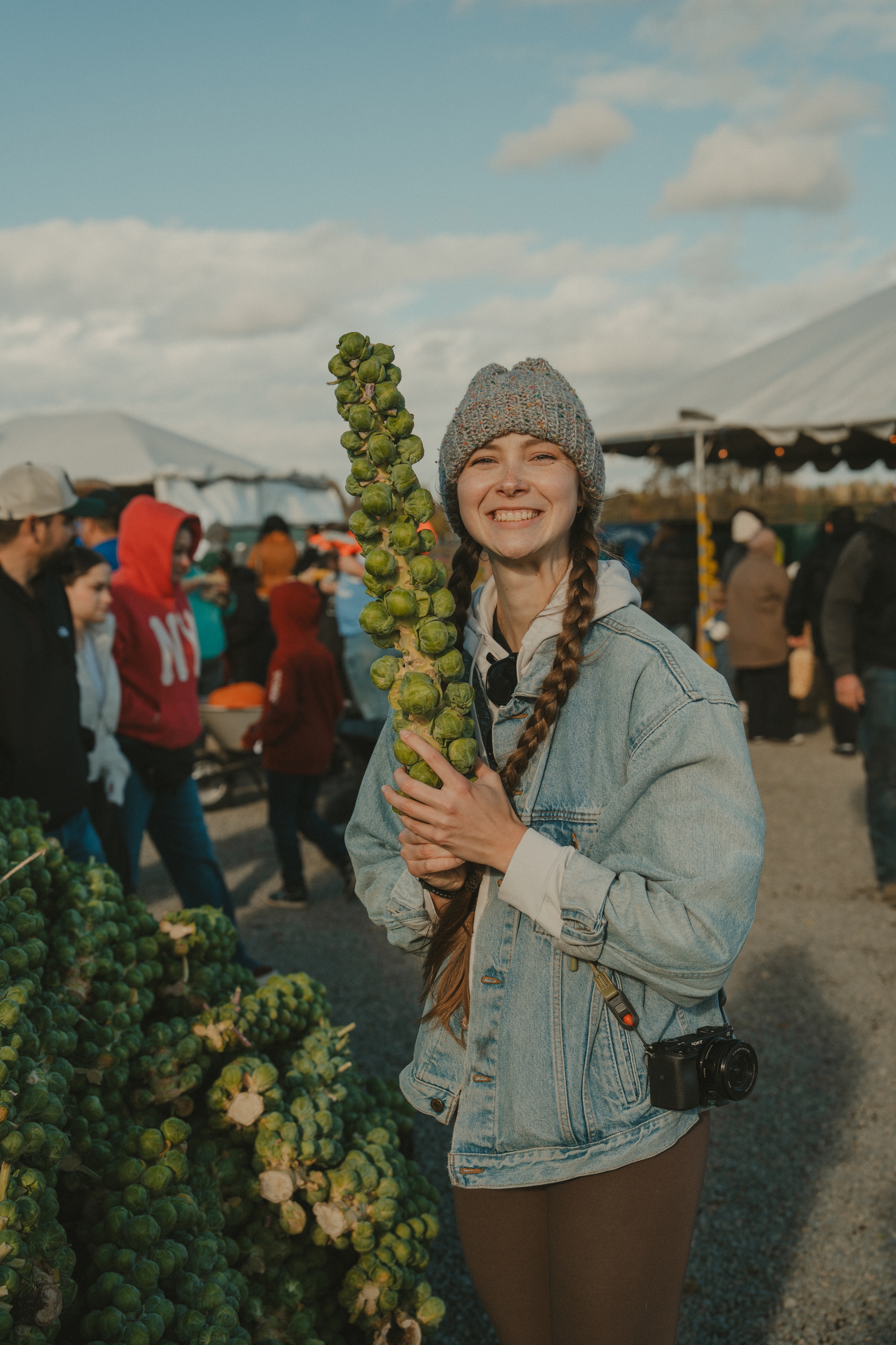 Person holding Brussels sprout stalk at a market, smiling with a crowd in the background