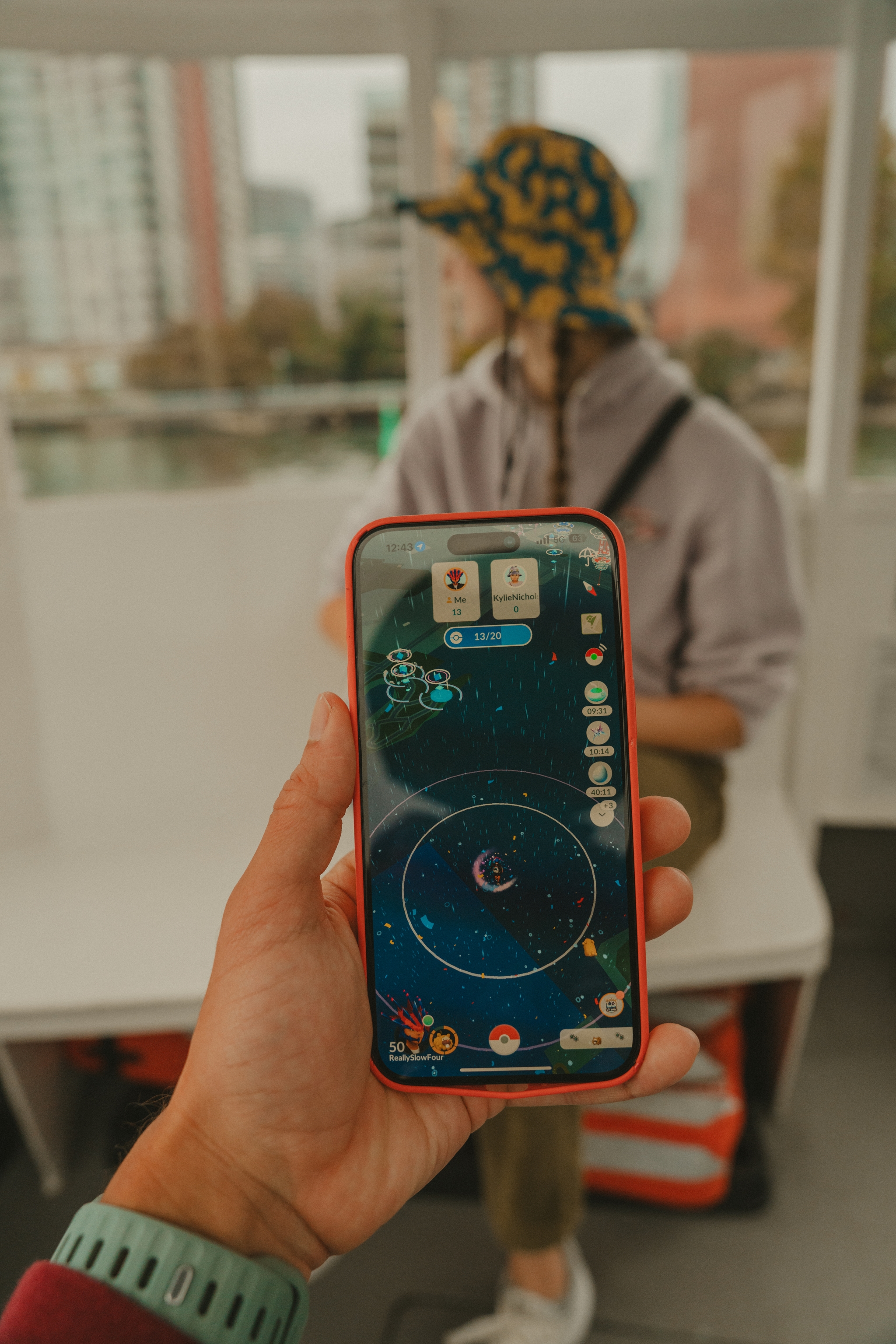 A person holds a smartphone displaying a game with a celestial theme, while in the background, another person sits in a casual setting, wearing a patterned bucket hat
