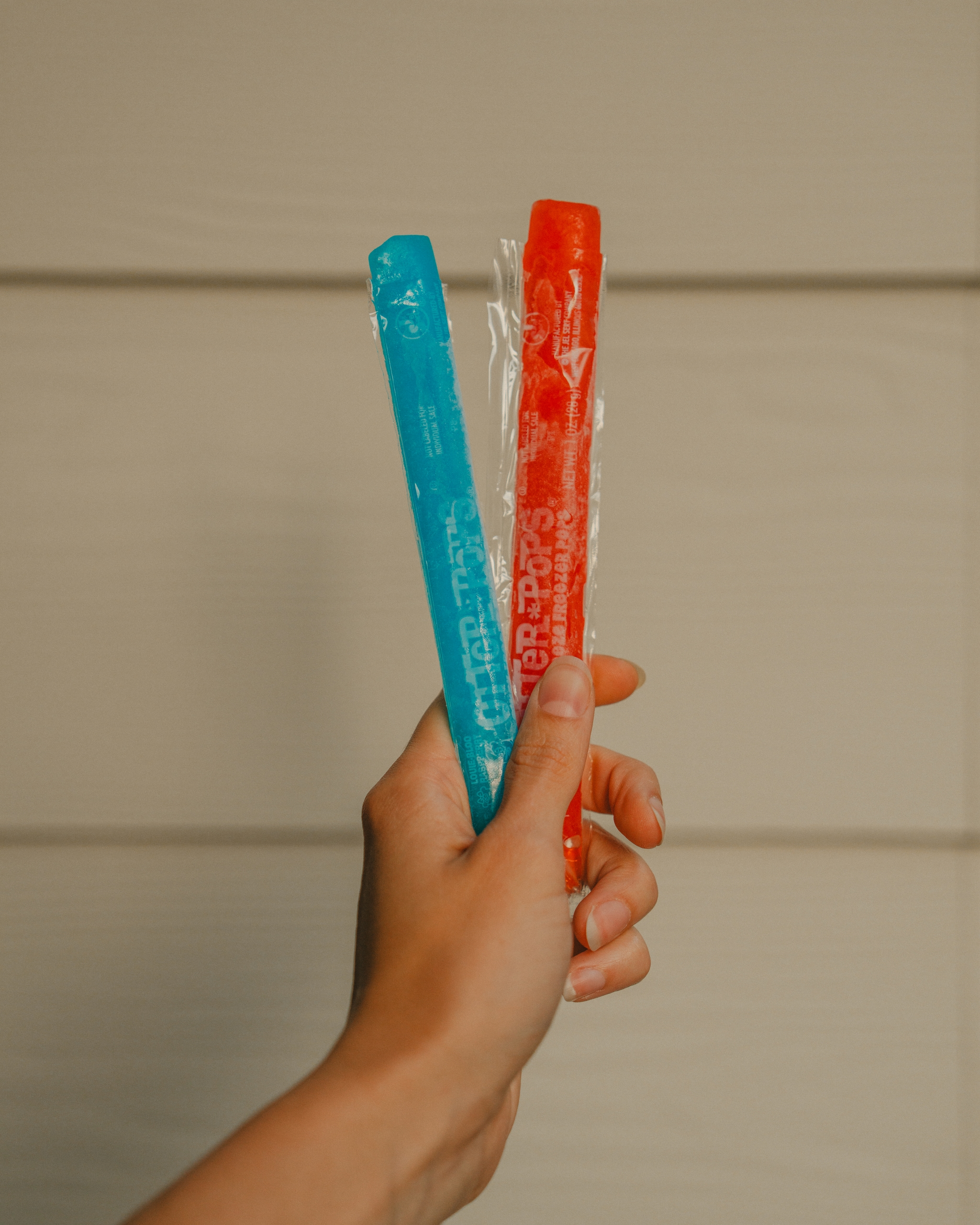 A hand holding two freeze pops, one blue and one red, against a neutral background