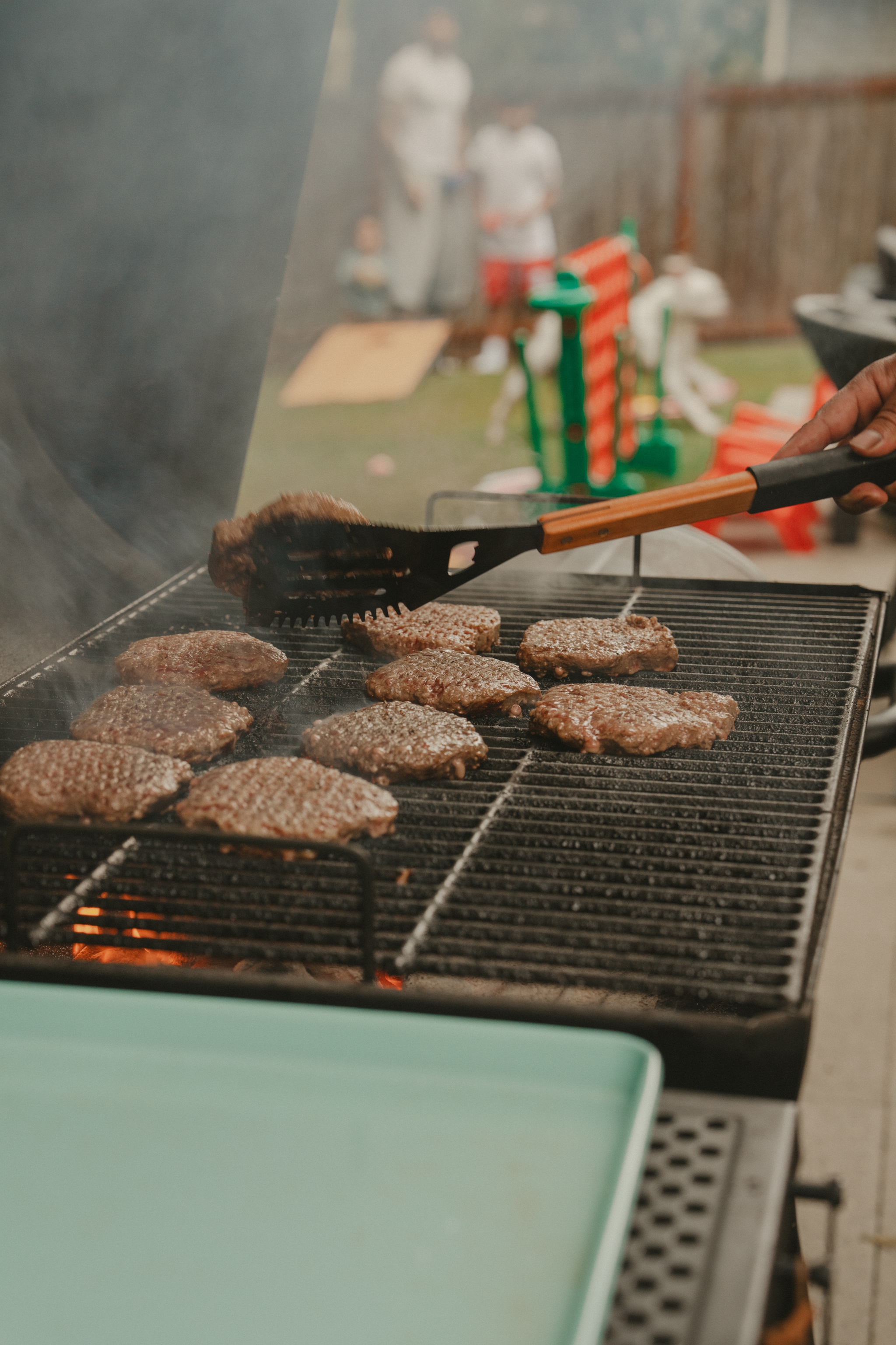 Grilling burgers on an outdoor barbecue, with smoke rising and people in the background