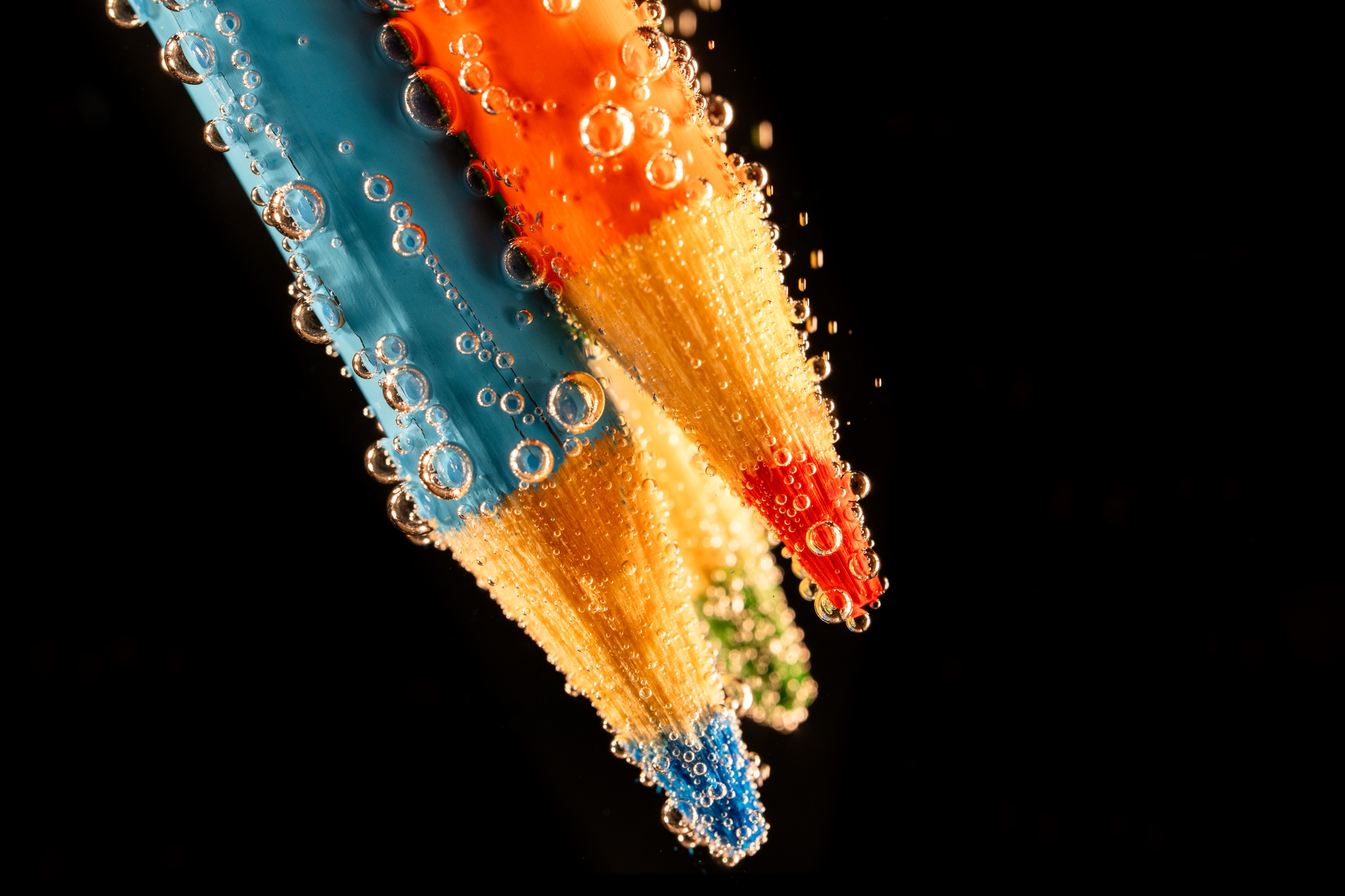 Three colored pencils (blue, orange, and red) submerged in water, covered with bubbles, against a black background