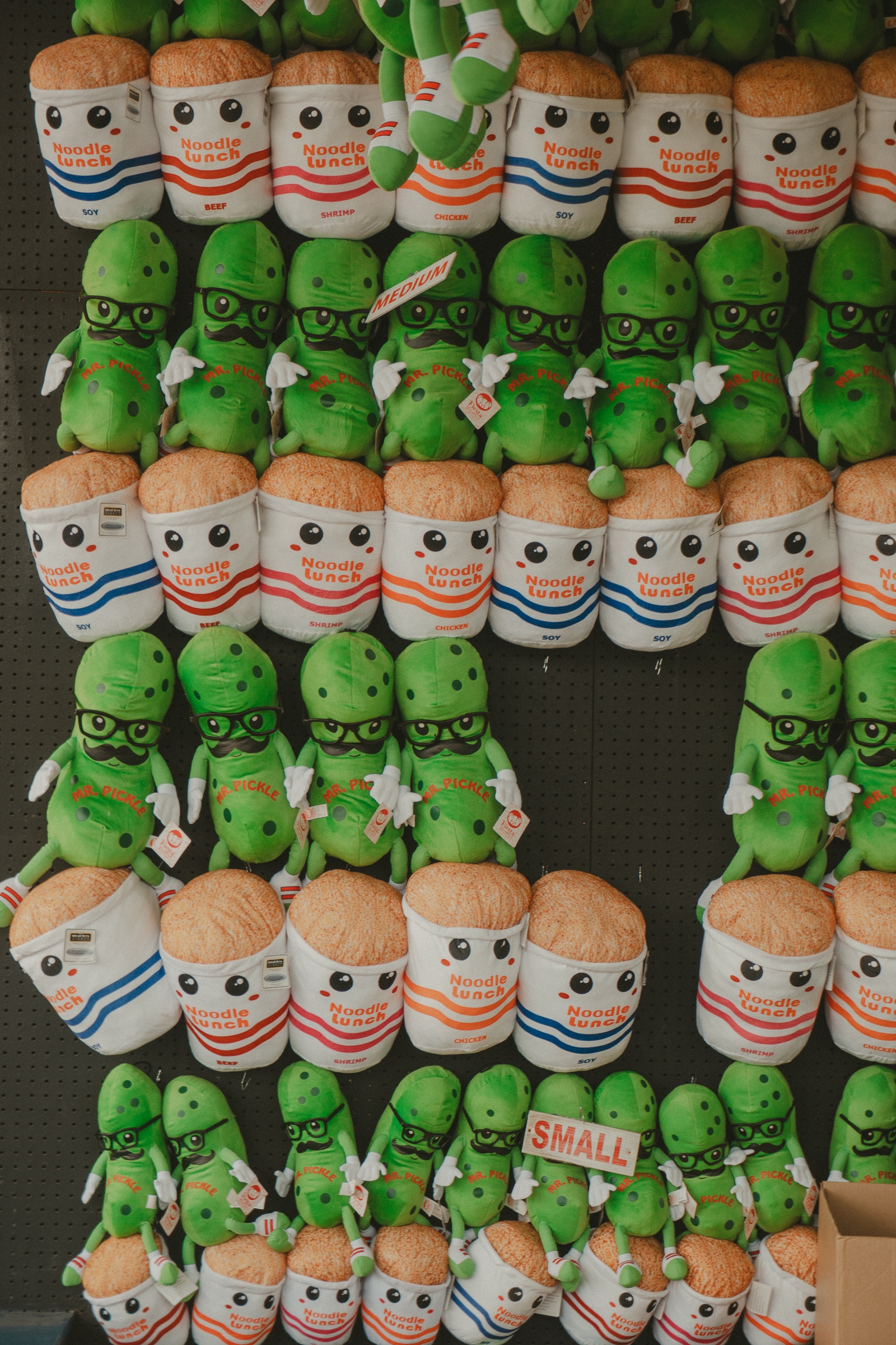 A display of plush toys, featuring rows of smiling green peas wearing glasses, and brown cylindrical shapes in striped shirts, arranged in alternating patterns
