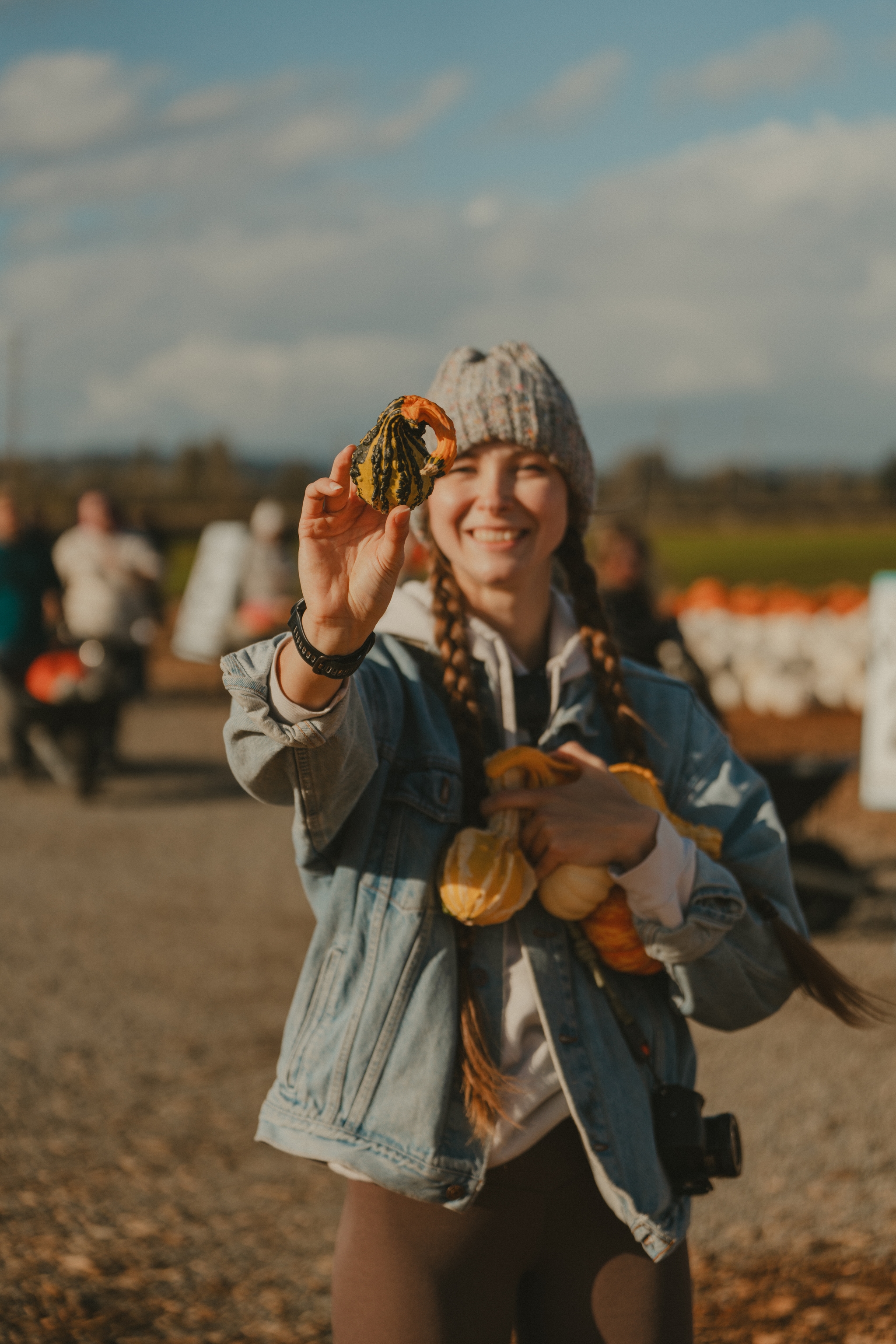 A person holding small pumpkins and gourds, wearing a beanie and denim jacket, with blurred background of a market or farm