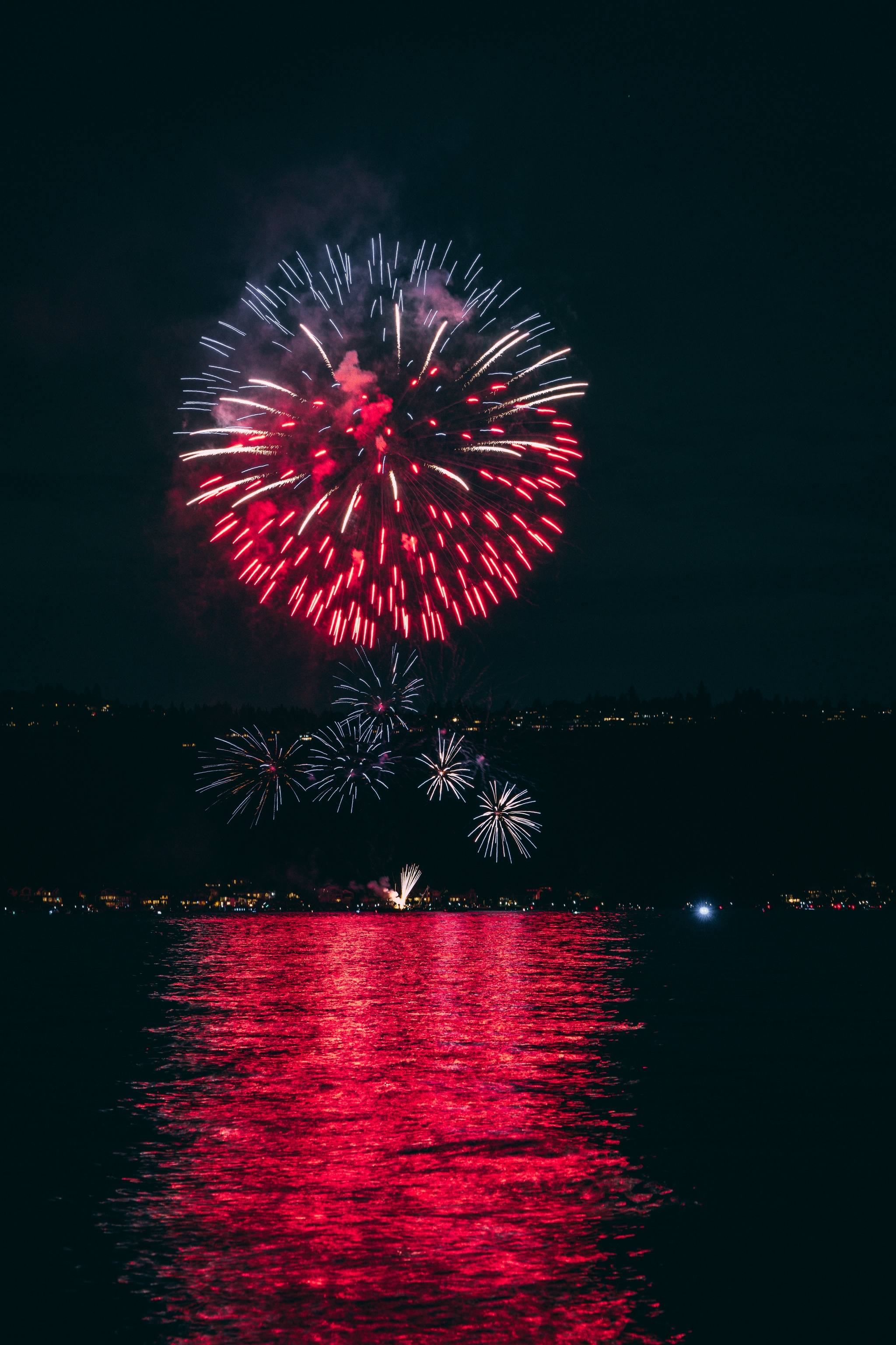 Fireworks exploding in the night sky over a body of water, with reflections of red and white lights on the surface