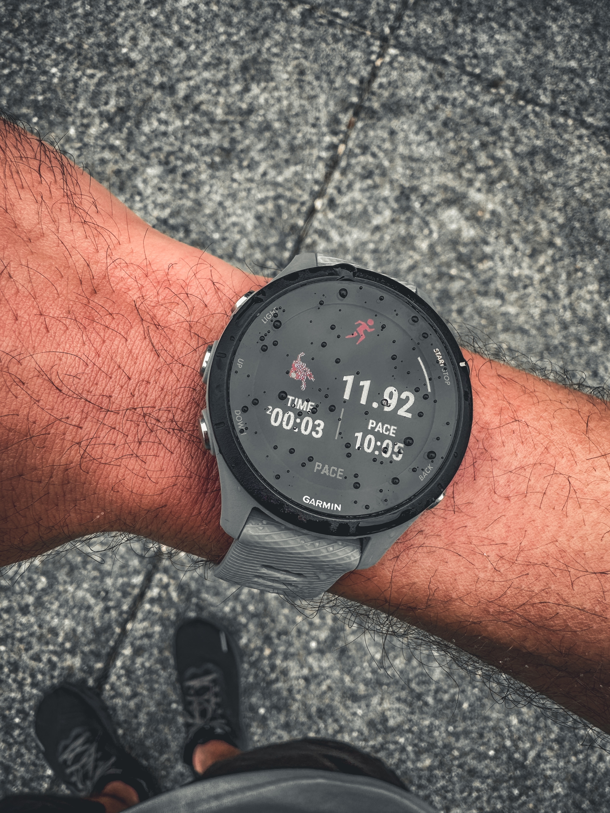 A person wearing a smartwatch displaying running statistics, including distance, pace, and time, with a textured ground surface in the background