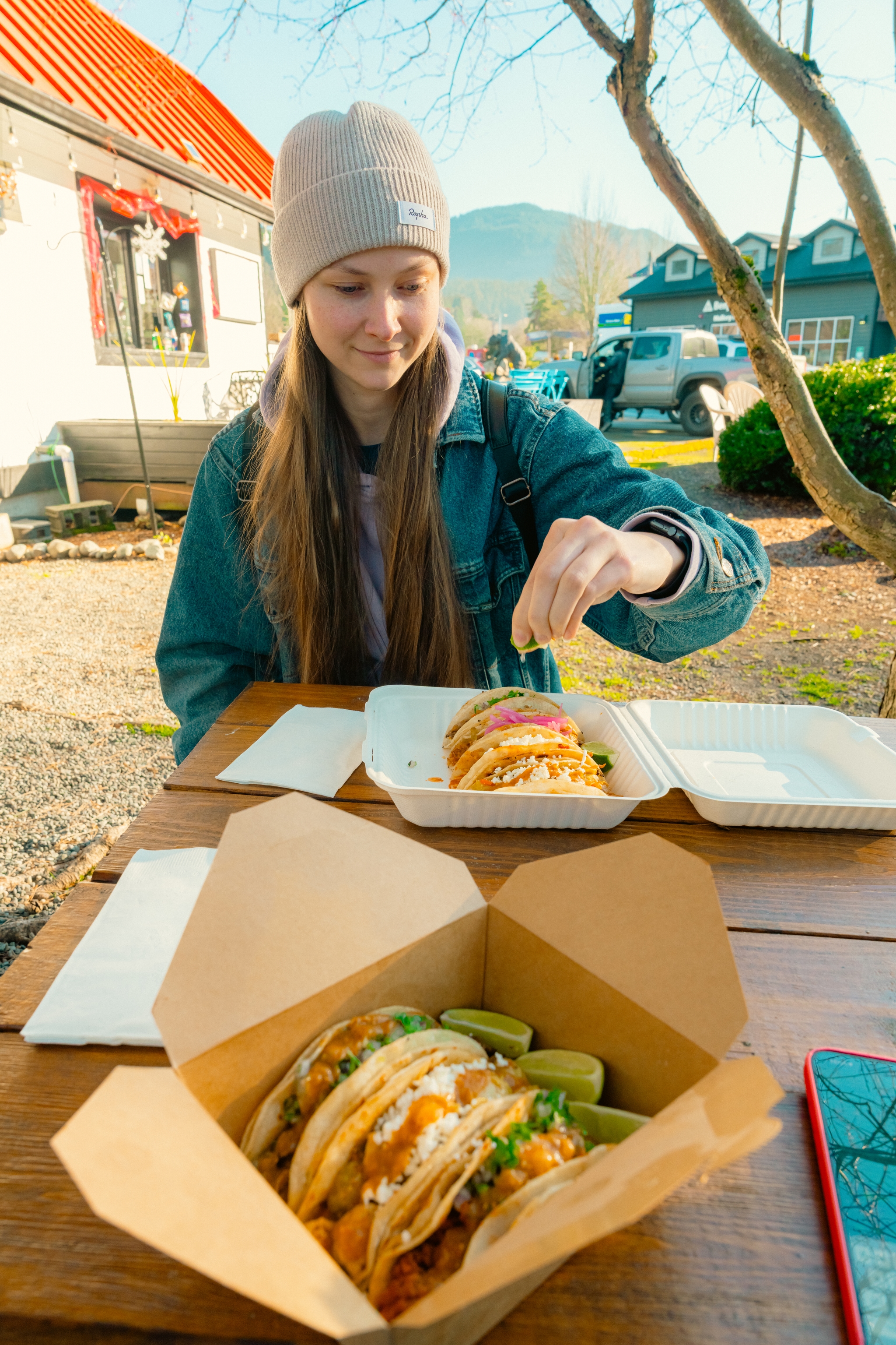 young woman eating takeout tacos at an outdoor wooden table with cardboard food containers and lime wedges in a casual restaurant setting