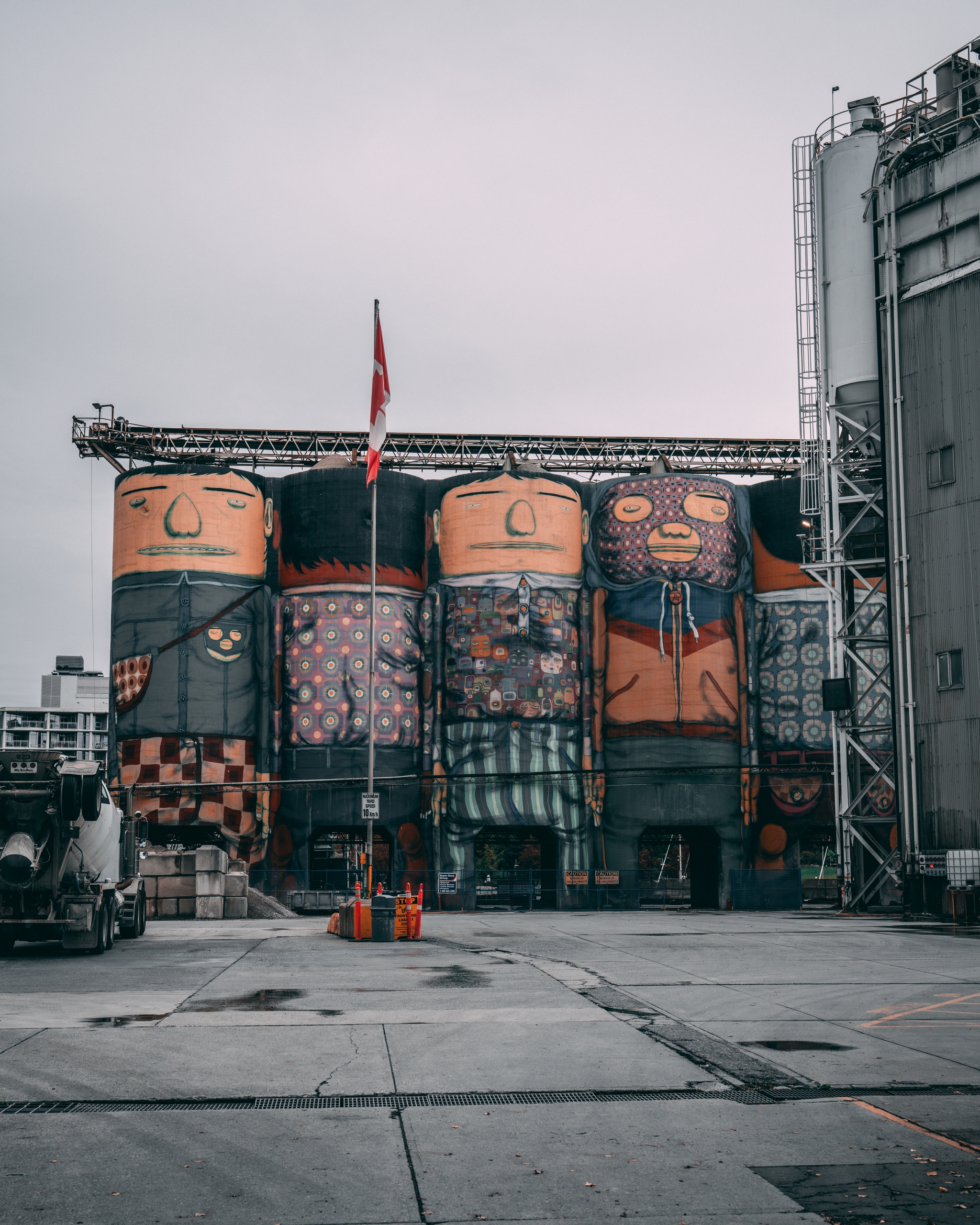 Large silos painted with colorful, abstract faces and patterns, set in an industrial area with a cloudy sky and a flag in the foreground