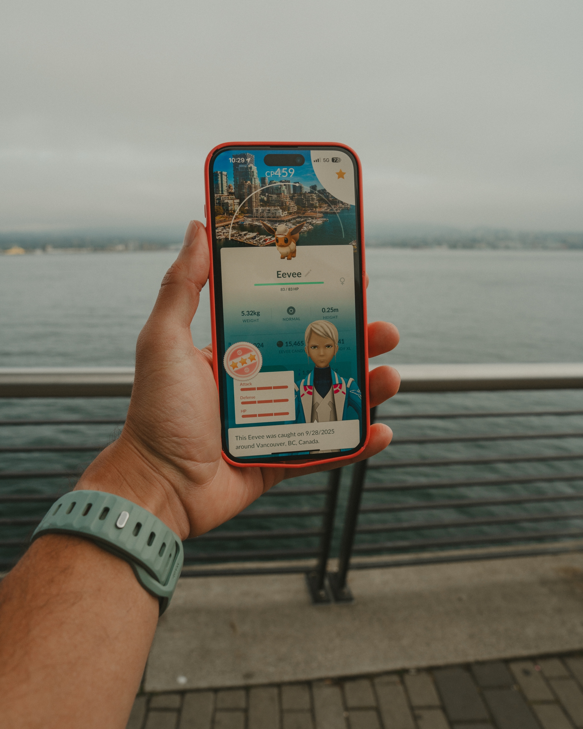 Hand holding a smartphone displaying a Pokémon screen, with waterfront railing and cloudy sky background