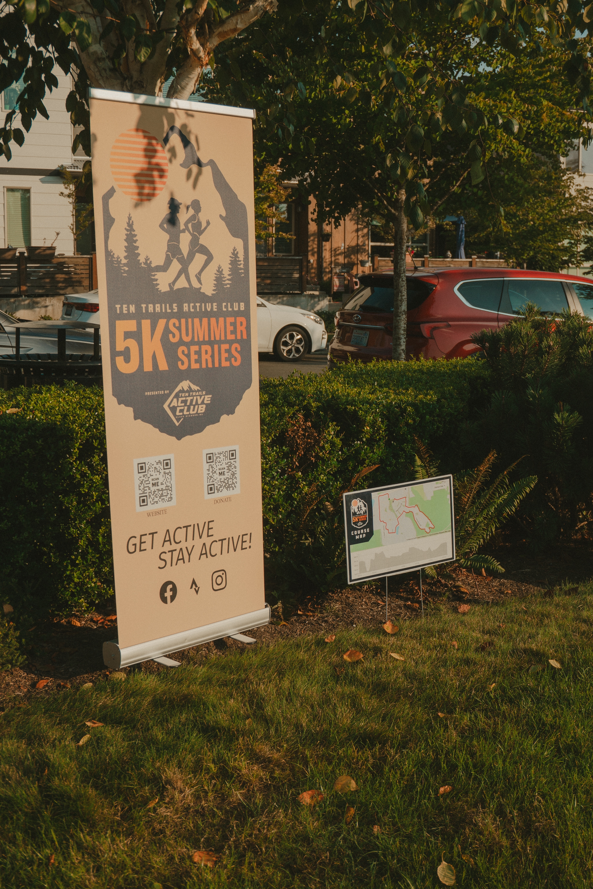 A banner advertising a 5K summer series event with the tagline Get Active, Stay Active stands on a grassy area next to a smaller sign with a map Cars and trees are in the background