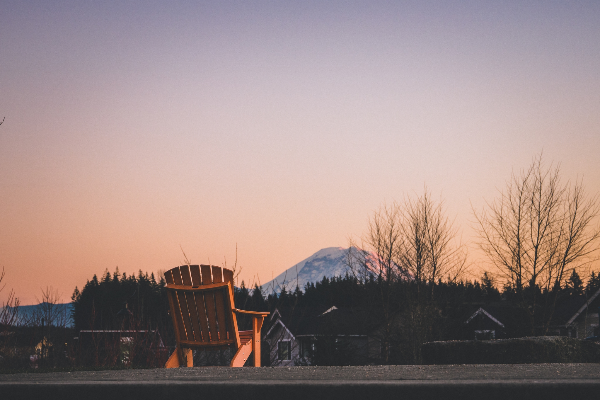 A wooden chair facing a snow-capped mountain at sunset, with bare trees and rooftops in the foreground