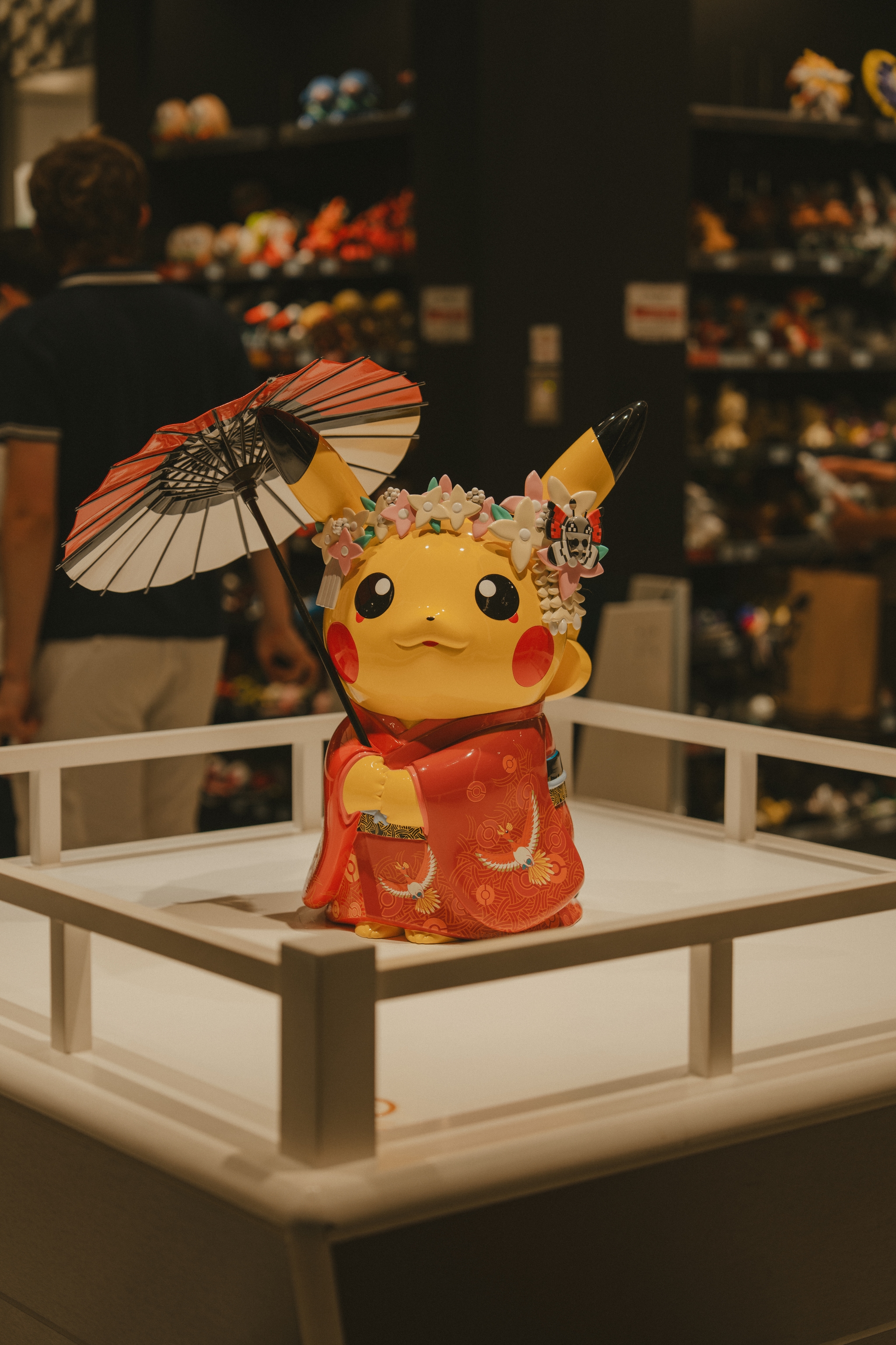 A Pikachu figurine dressed in a traditional Japanese kimono and holding a parasol, displayed on a platform in a store with various items in the background