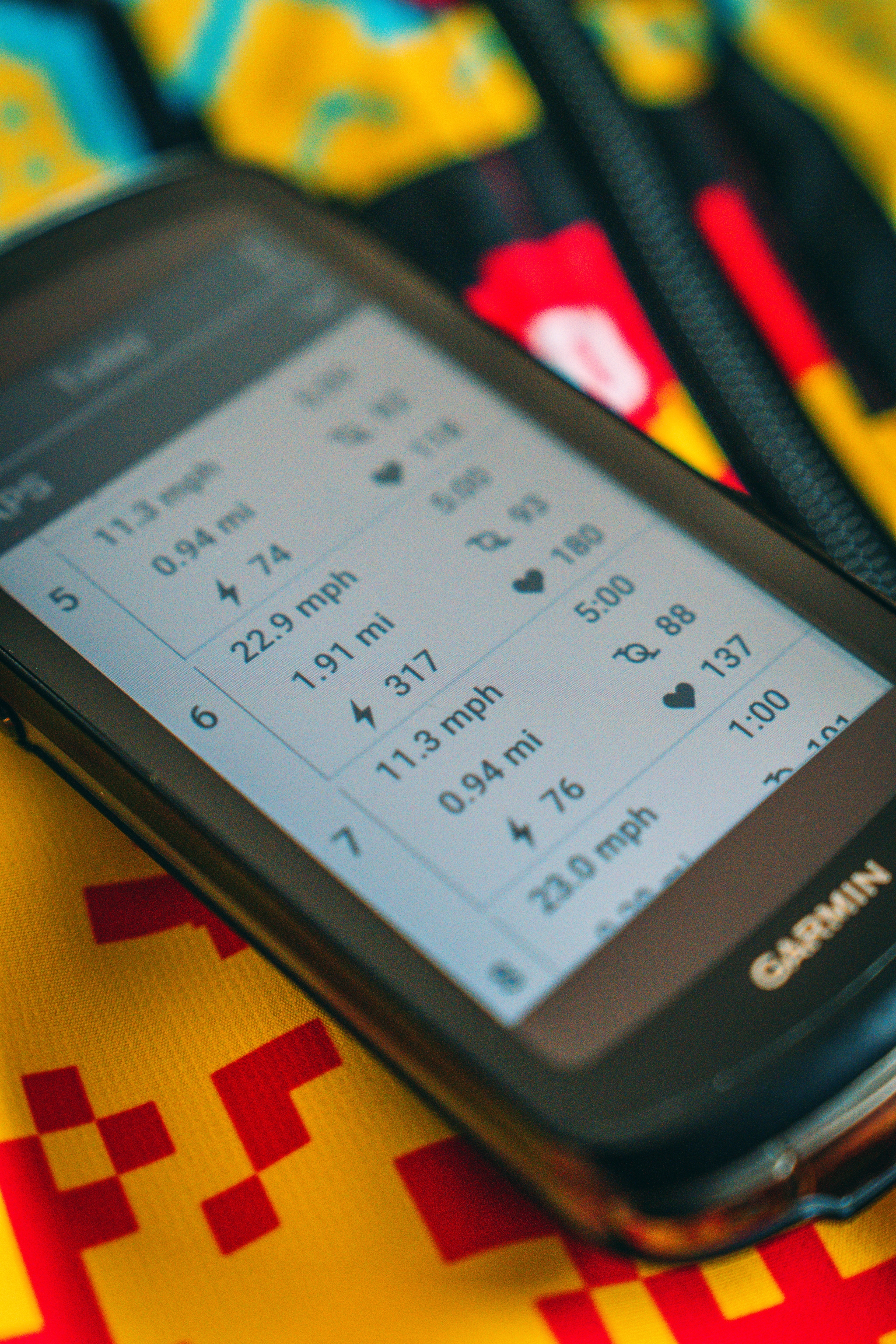A Garmin GPS device displays cycling statistics, including speed, distance, and heart rate data, resting on a colorful, patterned surface