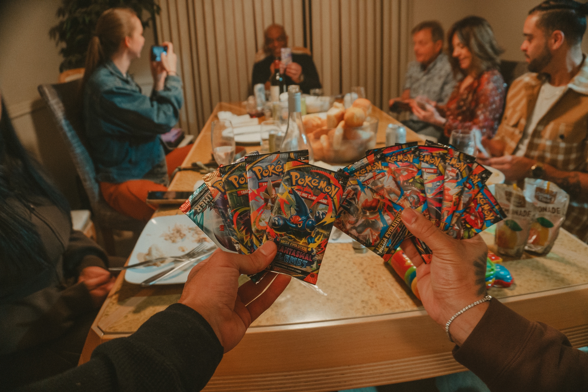 Hands holding trading cards at a dinner table with six people seated, suggesting a social gathering focused on card gameplay