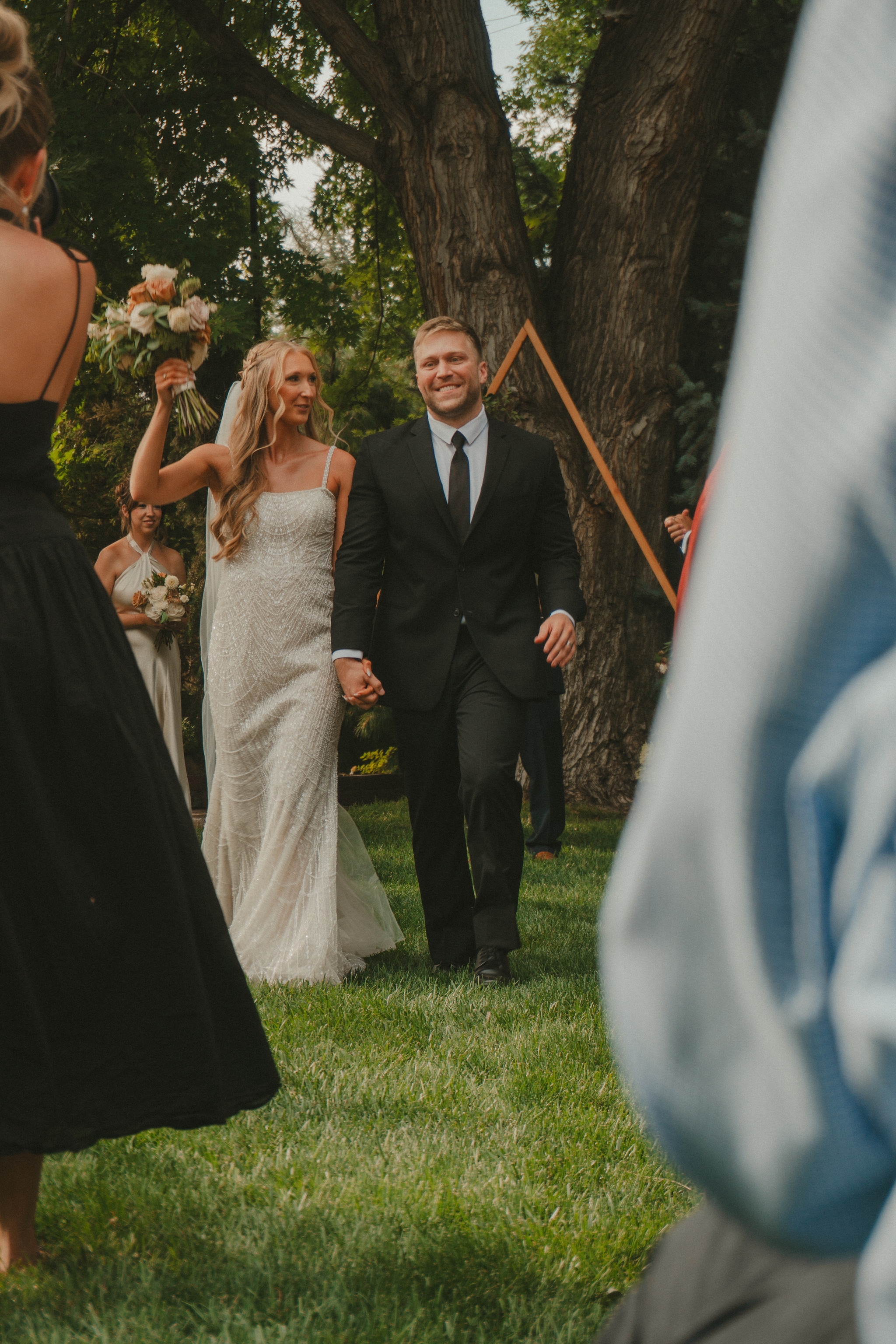 A bride and groom walk hand in hand outdoors, with the bride holding a bouquet and smiling They are surrounded by trees and guests, creating a joyful wedding atmosphere