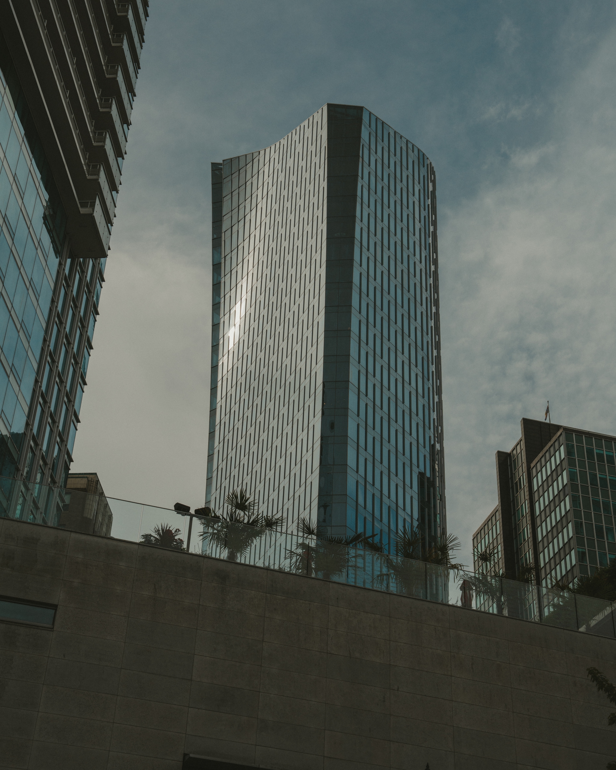 A modern skyscraper with reflective glass facade against a cloudy sky, surrounded by smaller buildings