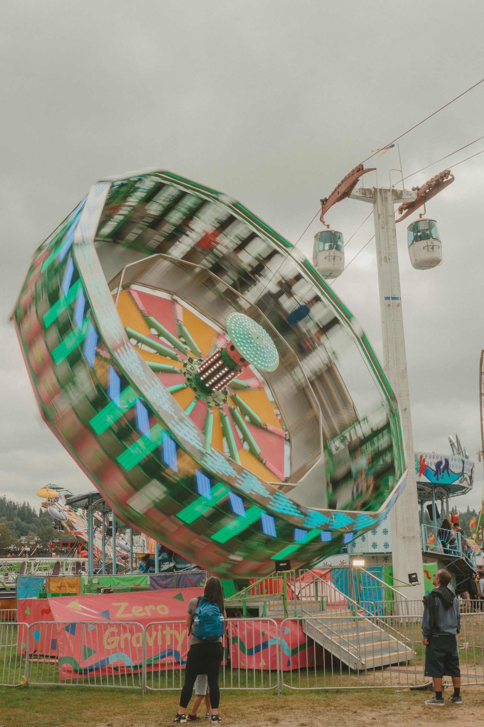 A colorful spinning amusement ride with lights, set against an overcast sky, alongside people standing and nearby gondola lift cabins