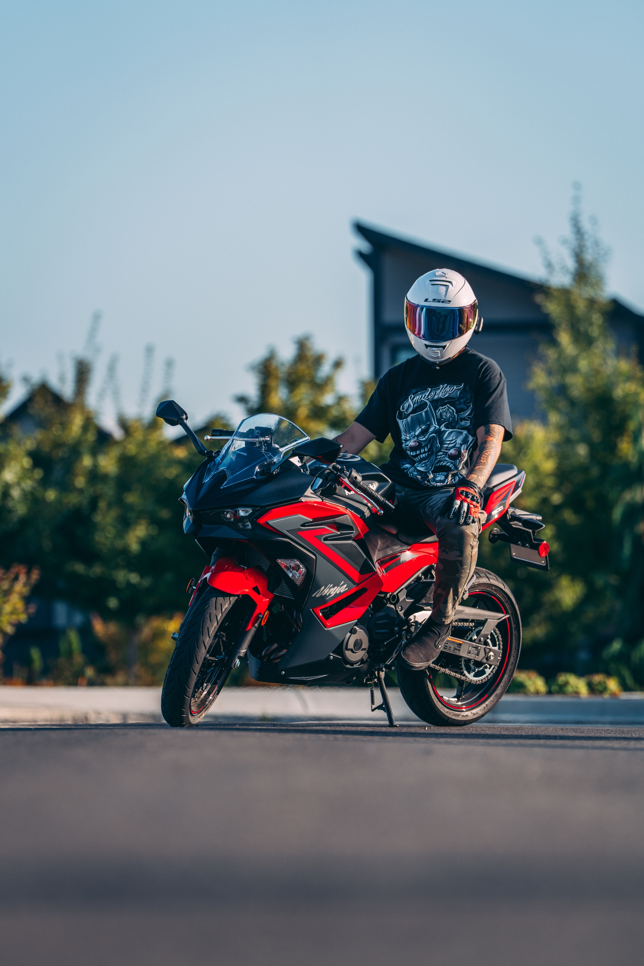 A person wearing a helmet and dark clothing sits on a red and black sport motorcycle on a paved surface with greenery in the background