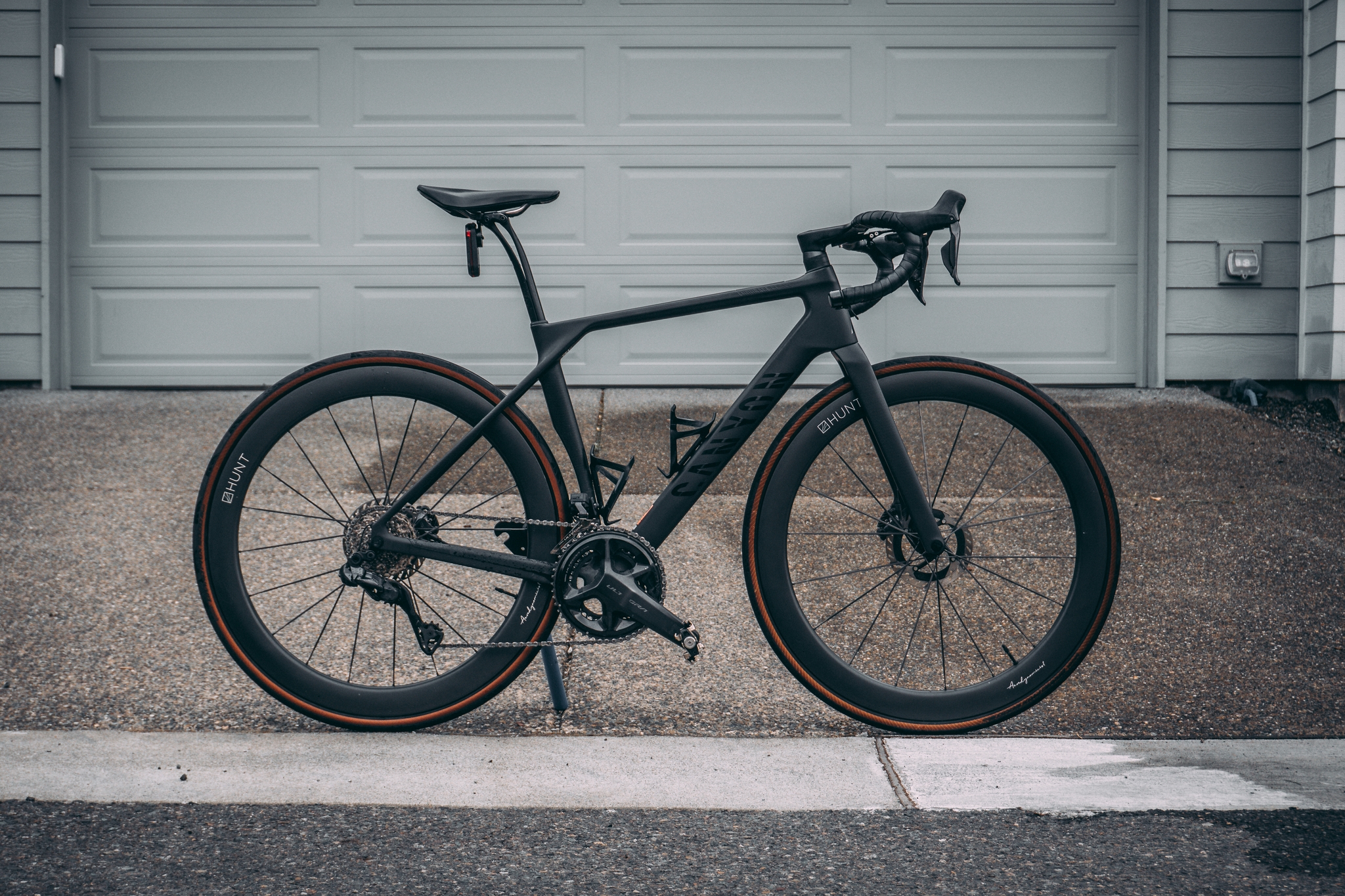 A sleek black road bike with drop handlebars and disc brakes, positioned in front of a closed garage door on a concrete driveway