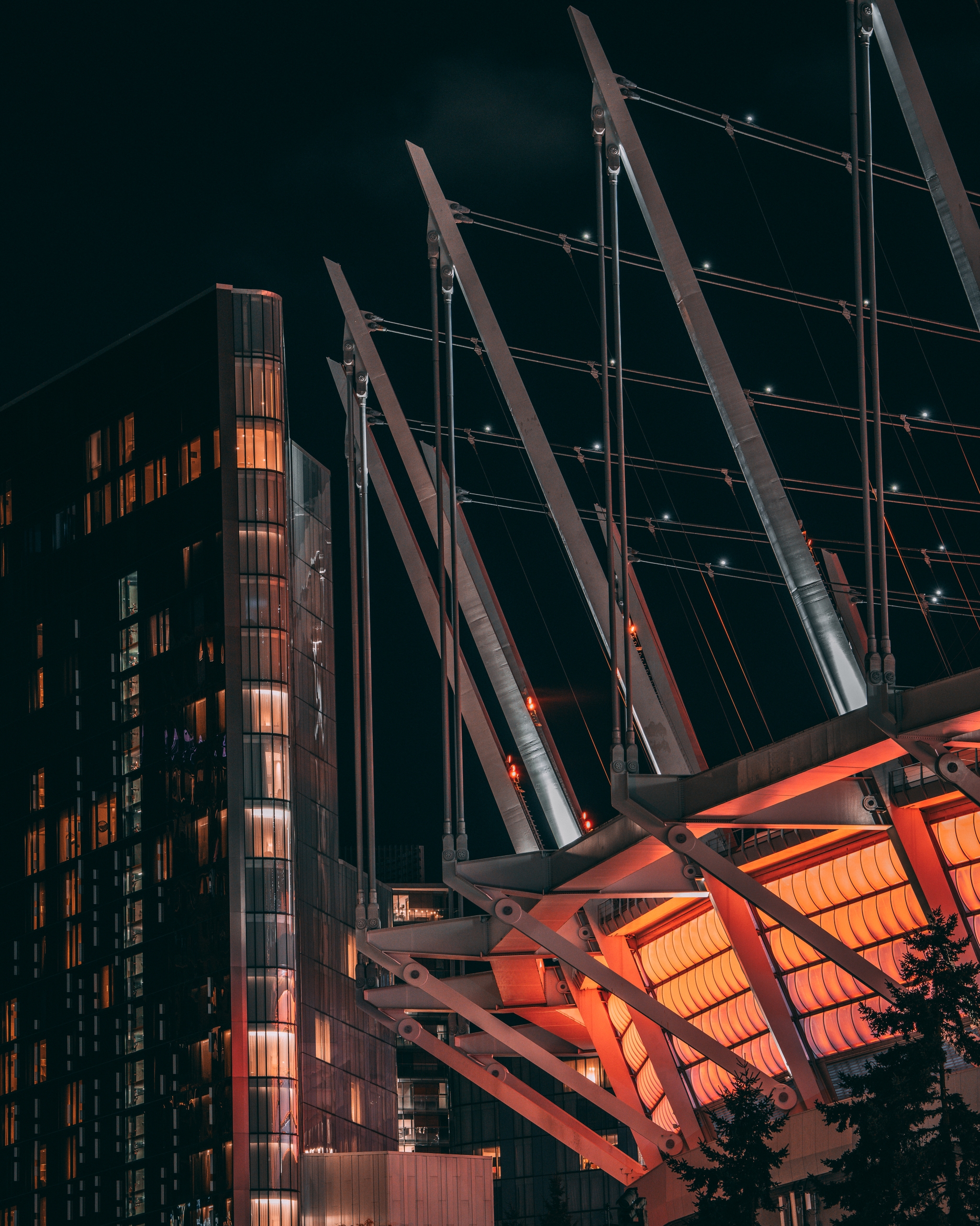 A modern architectural structure with angled beams and illuminated orange panels, set against a dark sky A nearby building with lit windows is visible