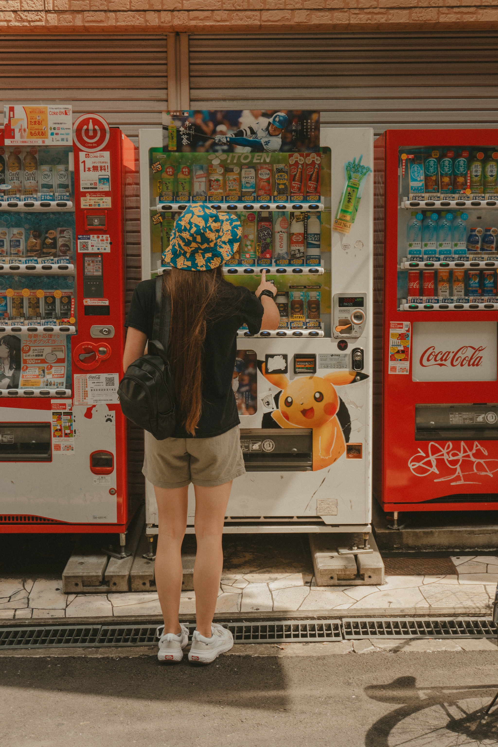 A person wearing a floral bucket hat and shorts is using a vending machine with a Pikachu design, flanked by two red vending machines