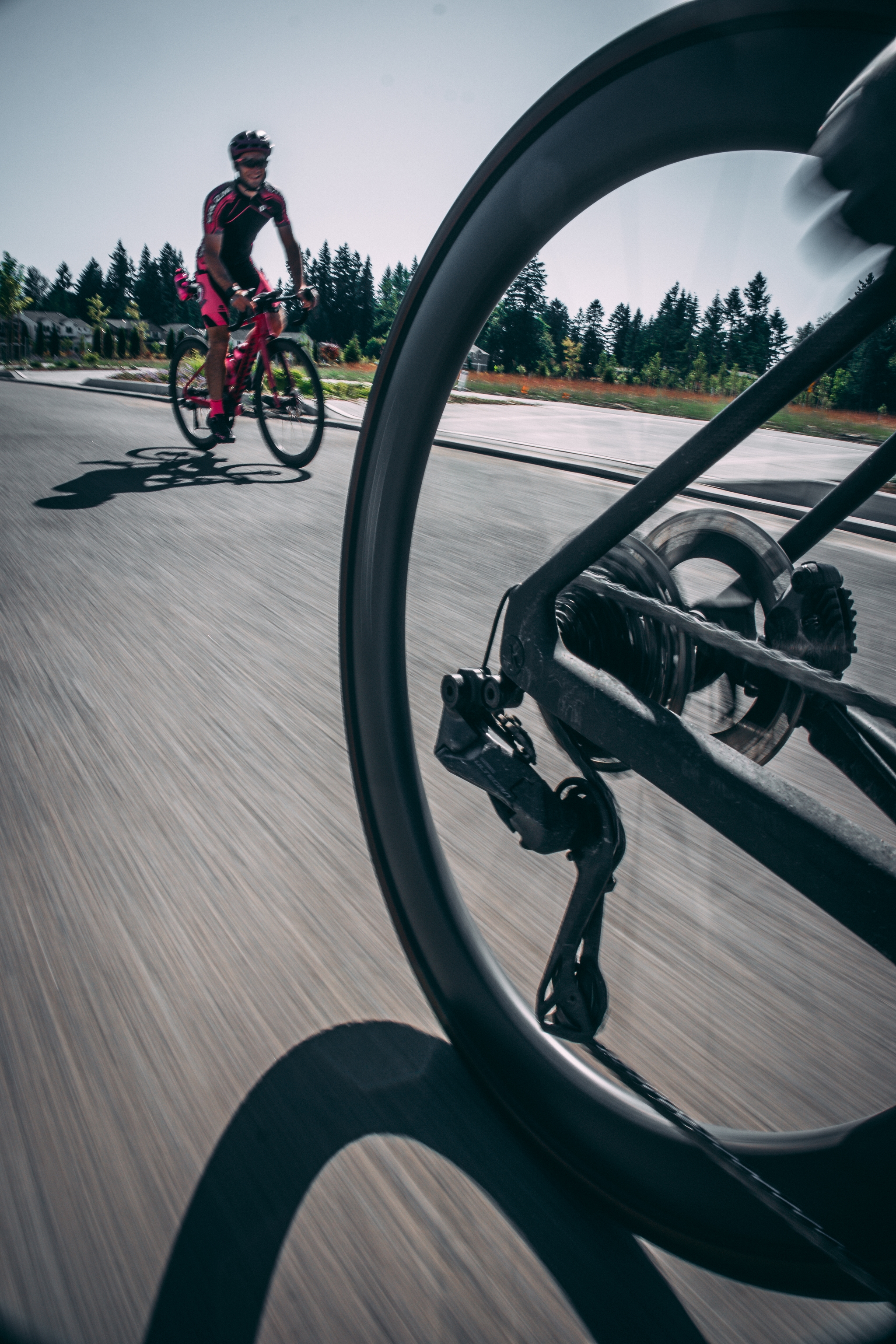 A cyclist in pink rides on a road, viewed through the spokes of another bicycle's wheel in the foreground