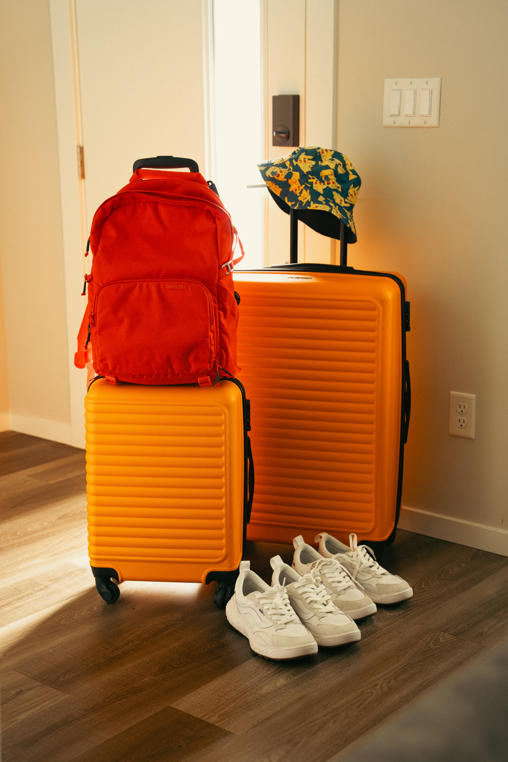 Two orange suitcases, a red backpack, a floral hat, and a pair of white sneakers are arranged on a wooden floor near a door