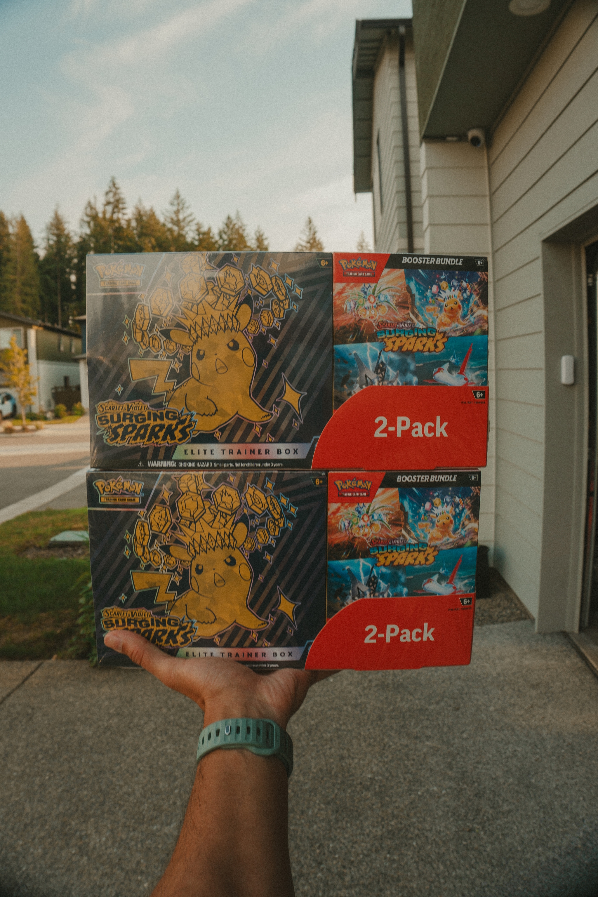 A hand holding two Pokémon card boxes labeled 2-Pack with colorful artwork, outside near a building