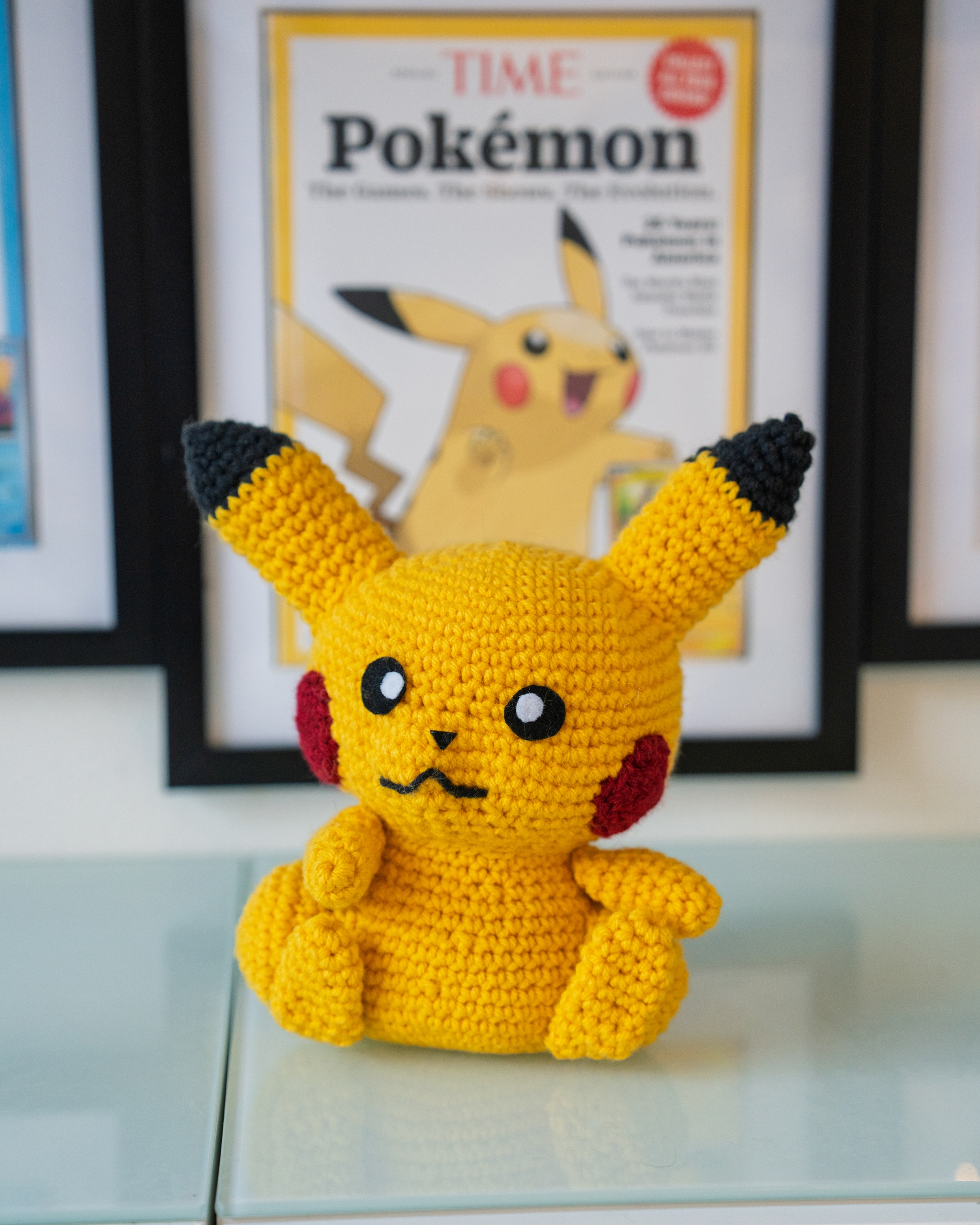 A crocheted Pikachu doll is placed in front of a framed Pokémon-themed magazine cover