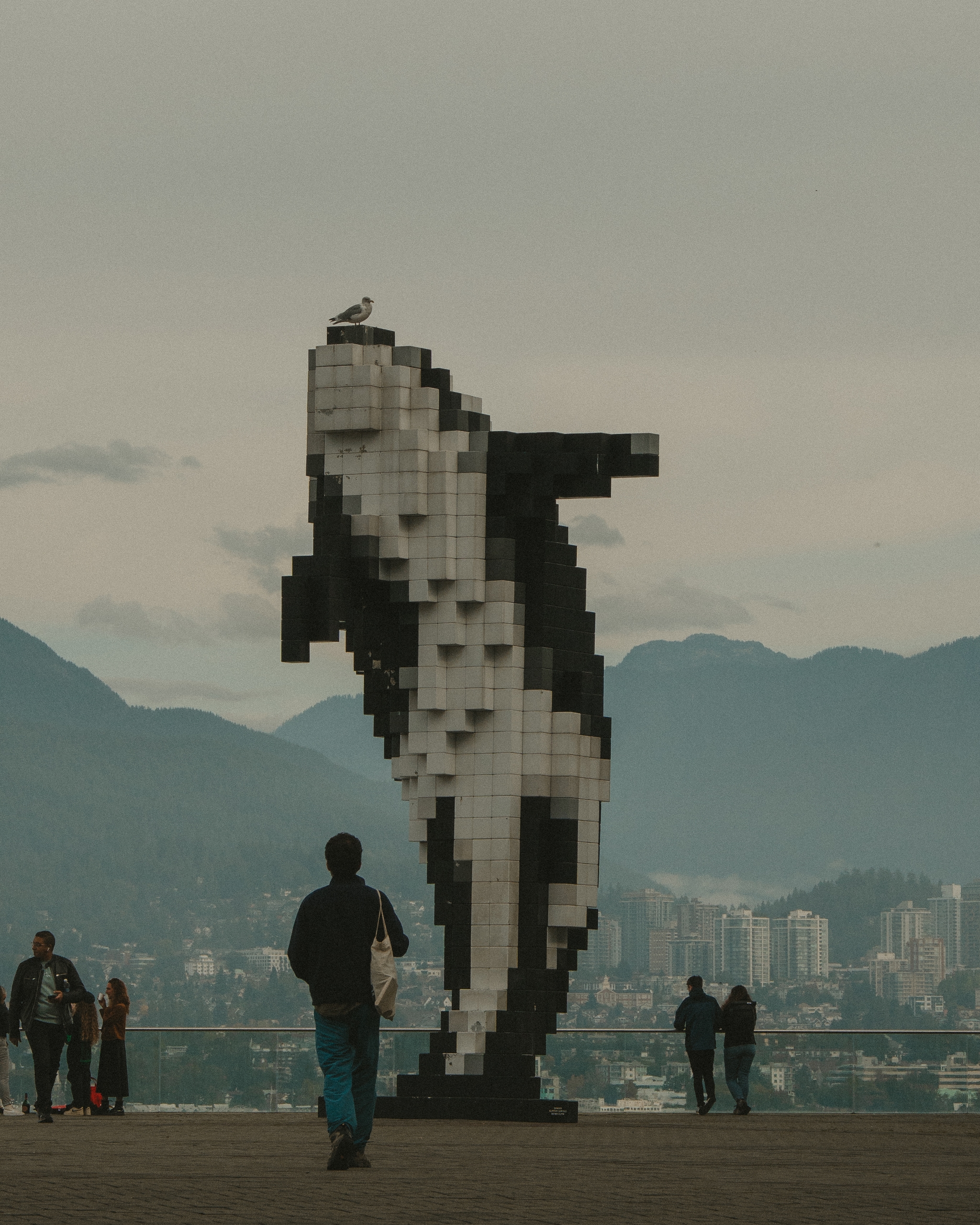 A pixelated orca sculpture stands prominently in an outdoor setting with people walking nearby and a cityscape with mountains in the background