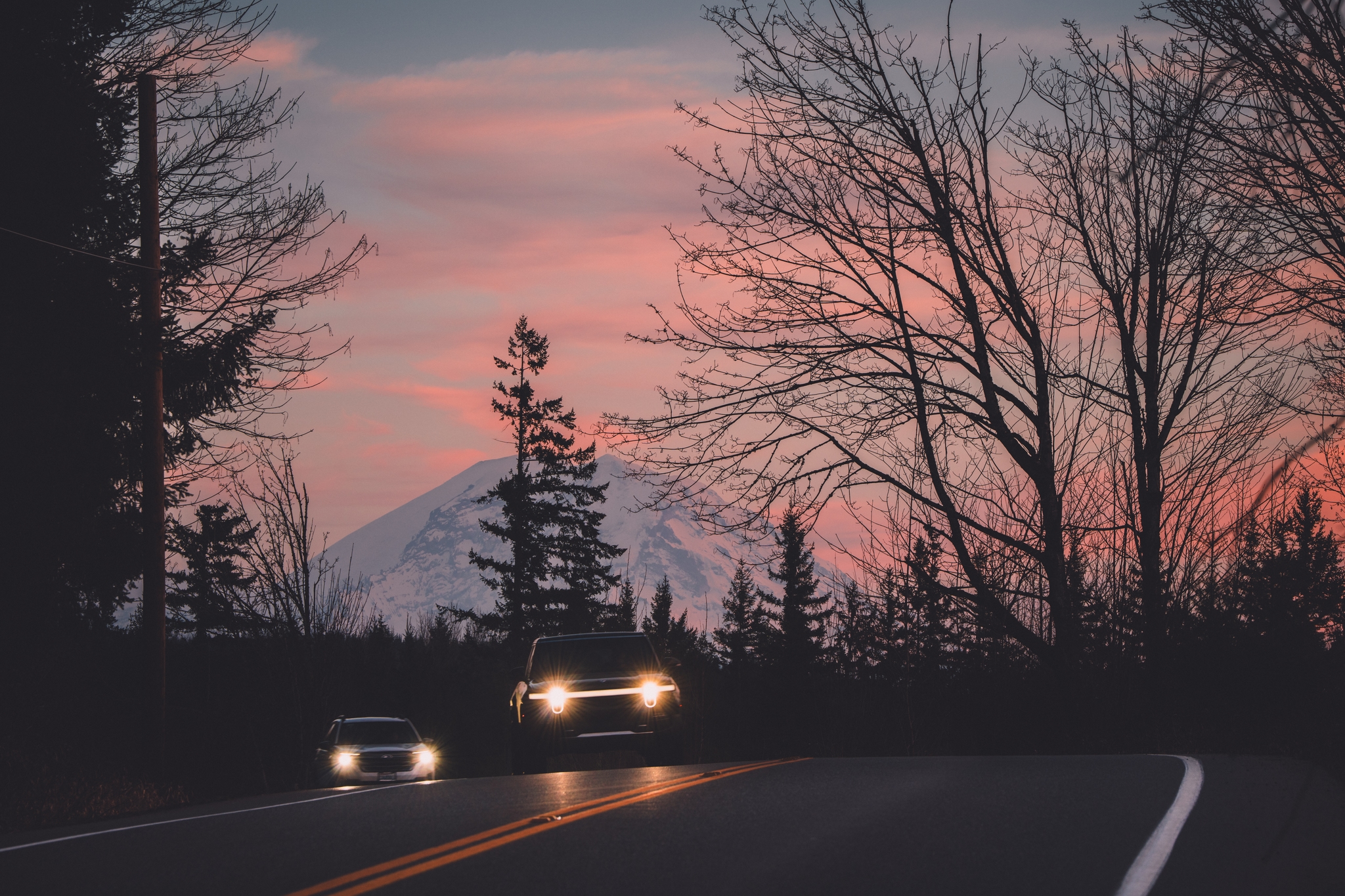 A scenic road at dusk with cars approaching, framed by silhouetted trees and a snow-capped mountain in the background under a pink and blue sky