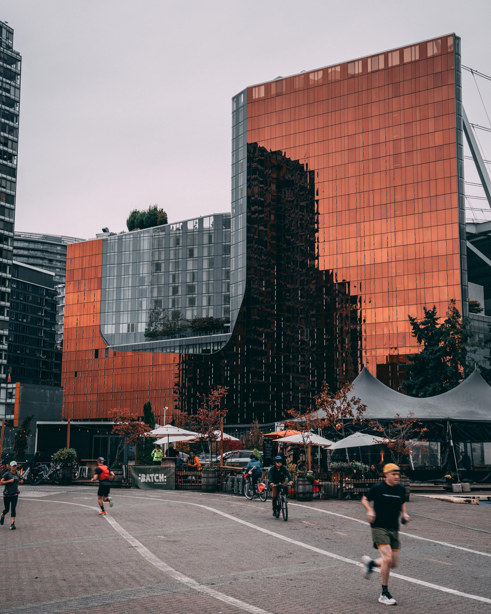 A modern cityscape featuring a reflective, copper-colored building with people jogging and cycling in the foreground