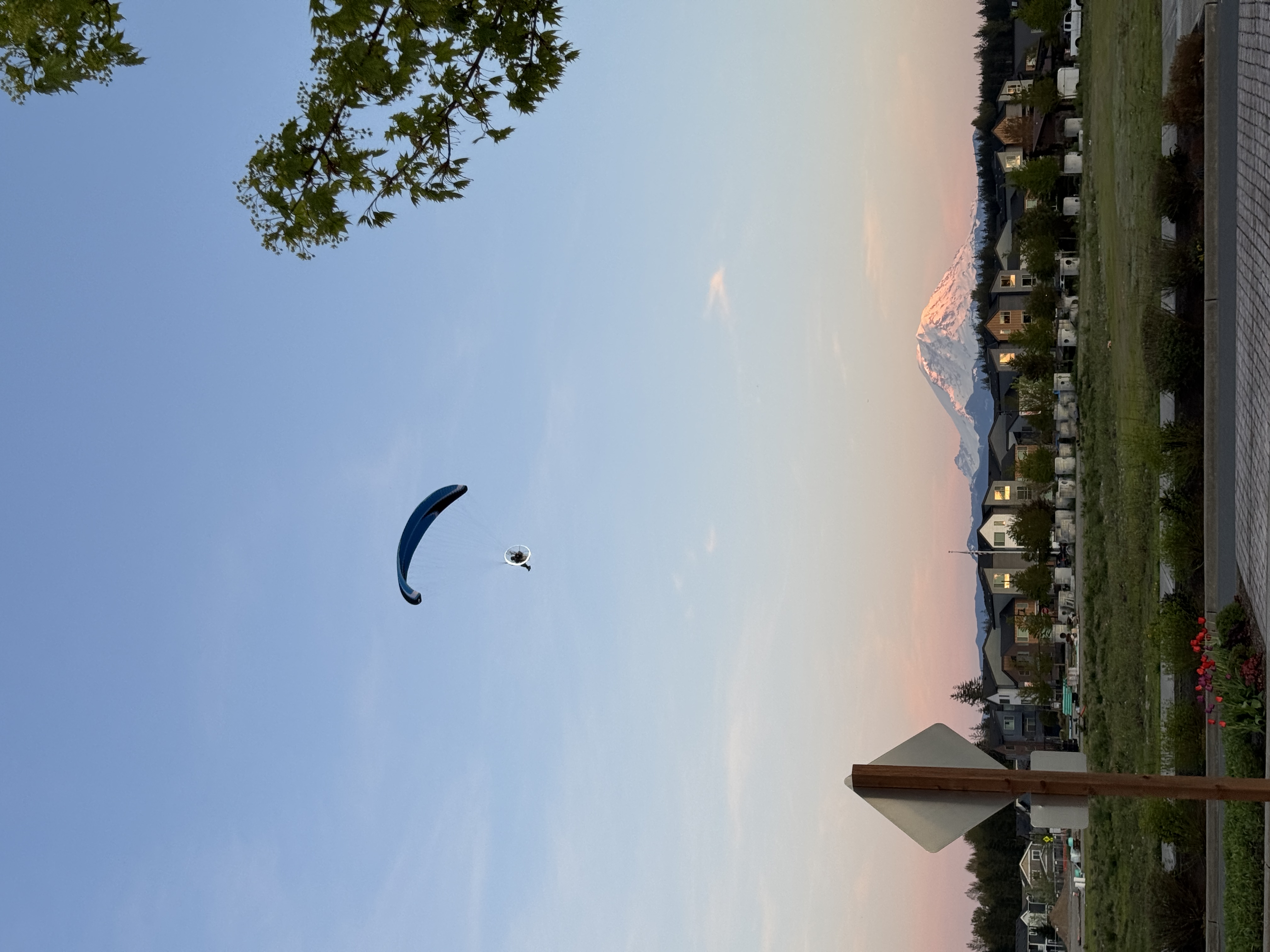 A paraglider is soaring in the sky above a suburban area with a snow-capped mountain in the background during sunset