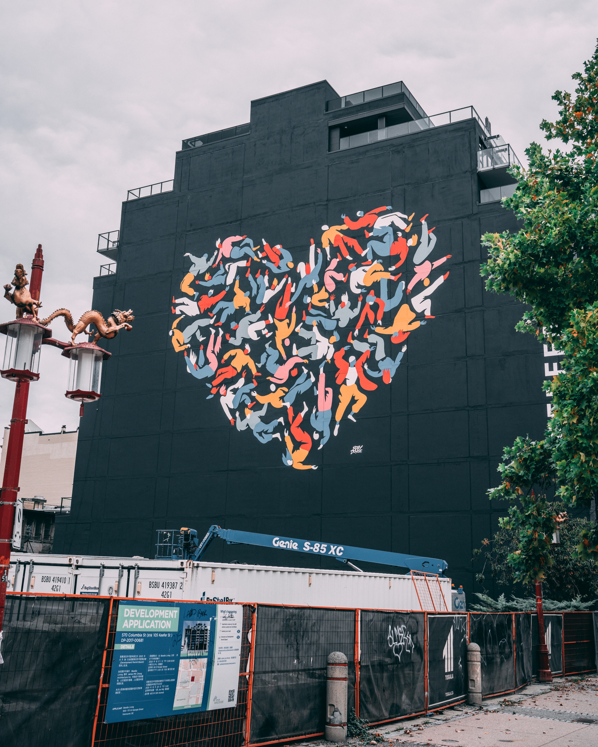A large mural featuring a colorful heart composed of various abstract shapes is painted on the side of a tall, dark building. A decorative street lamp and some trees are in the foreground