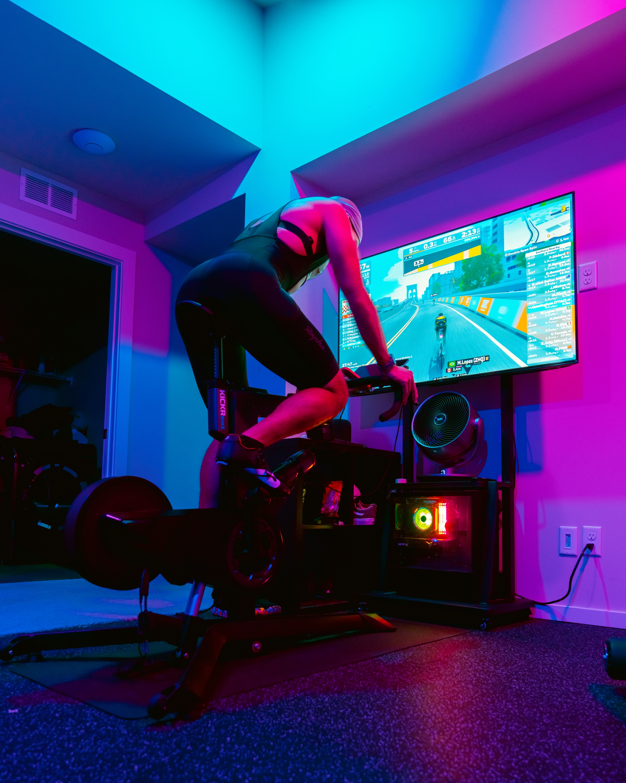 Person cycling on an indoor bike with a virtual cycling game displayed on a screen, illuminated by neon lights