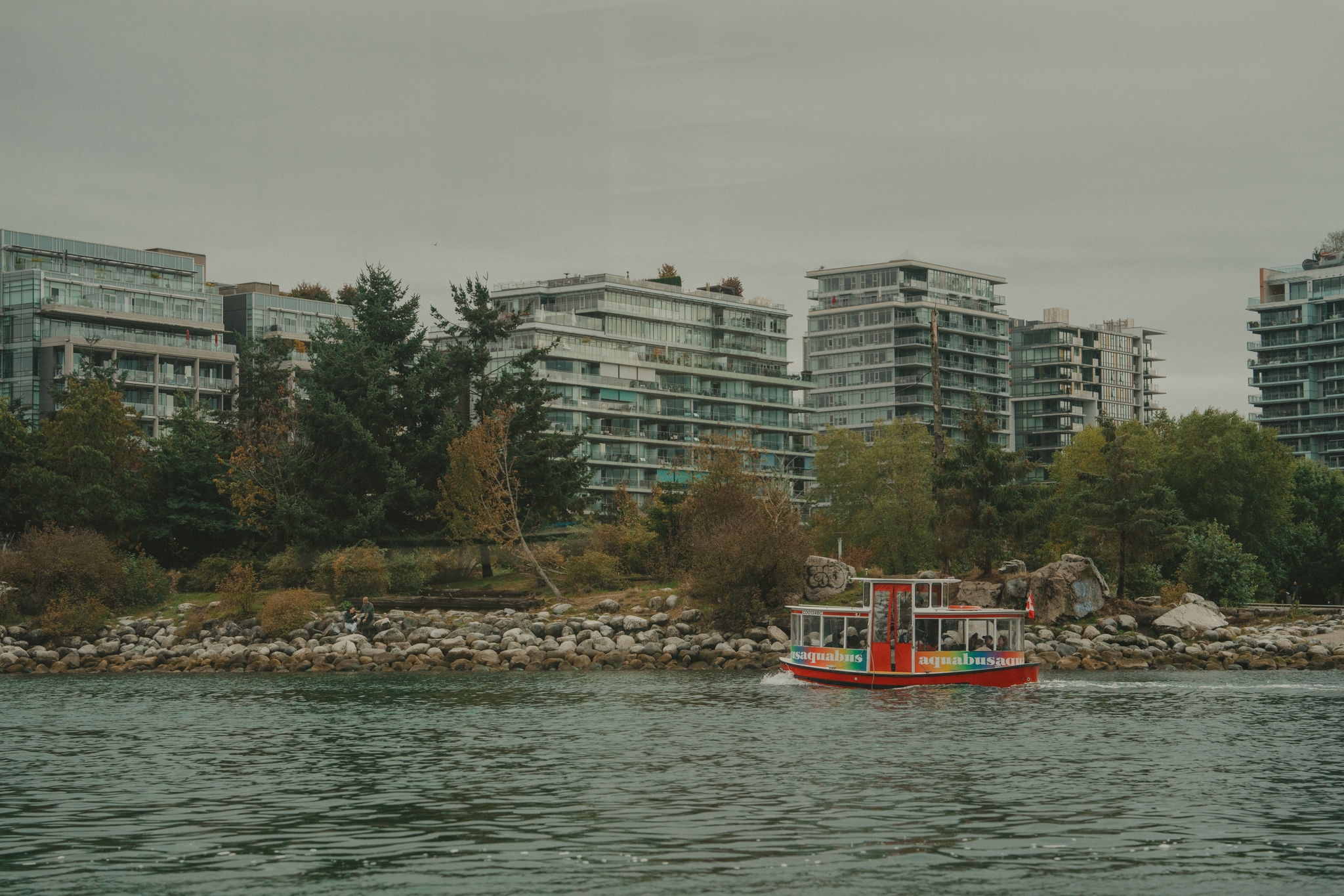 A small red and yellow boat is on the water near a rocky shoreline with apartment buildings and trees in the background. The sky is overcast