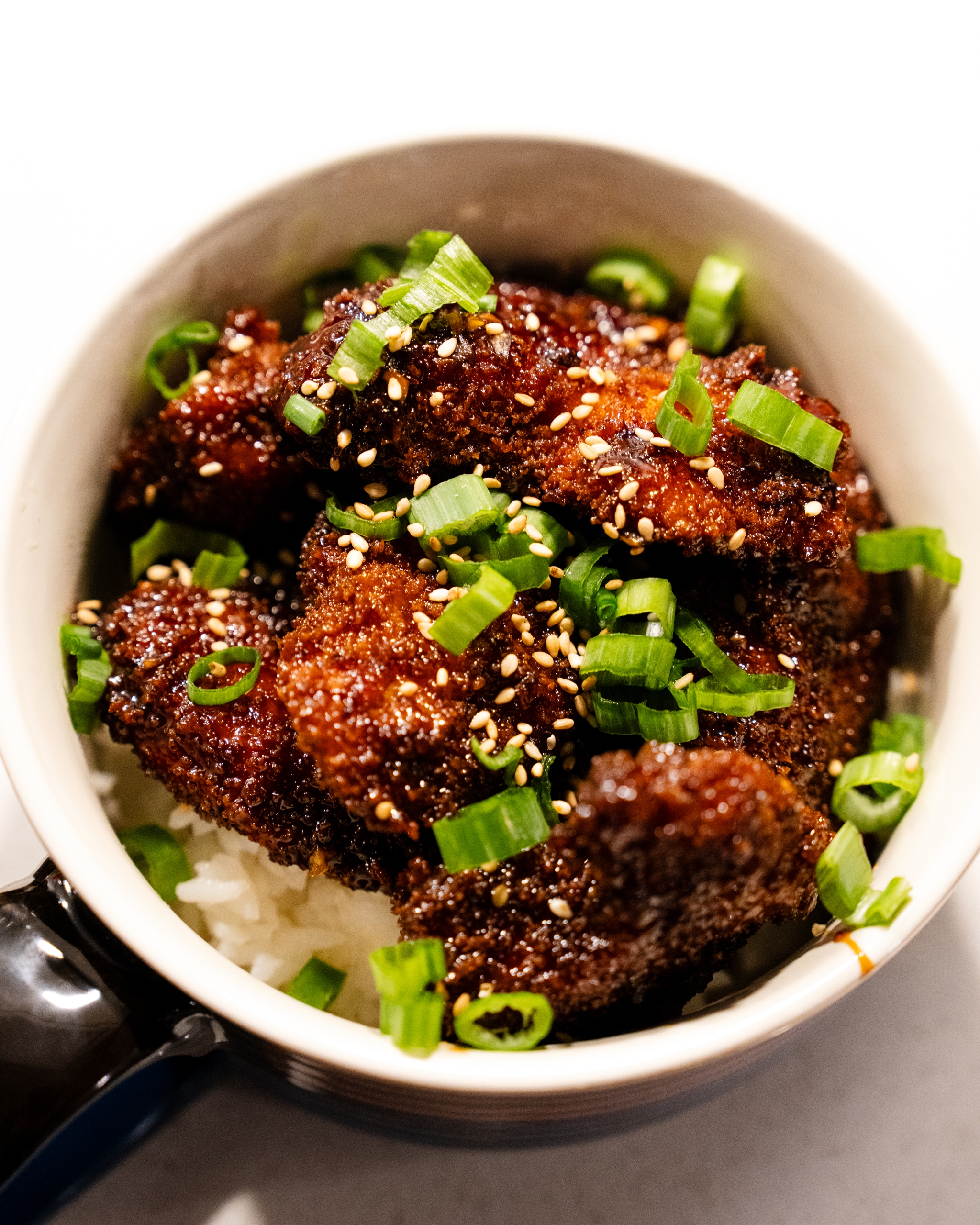 A bowl of crispy coated food topped with green scallions and sesame seeds, served over a bed of rice