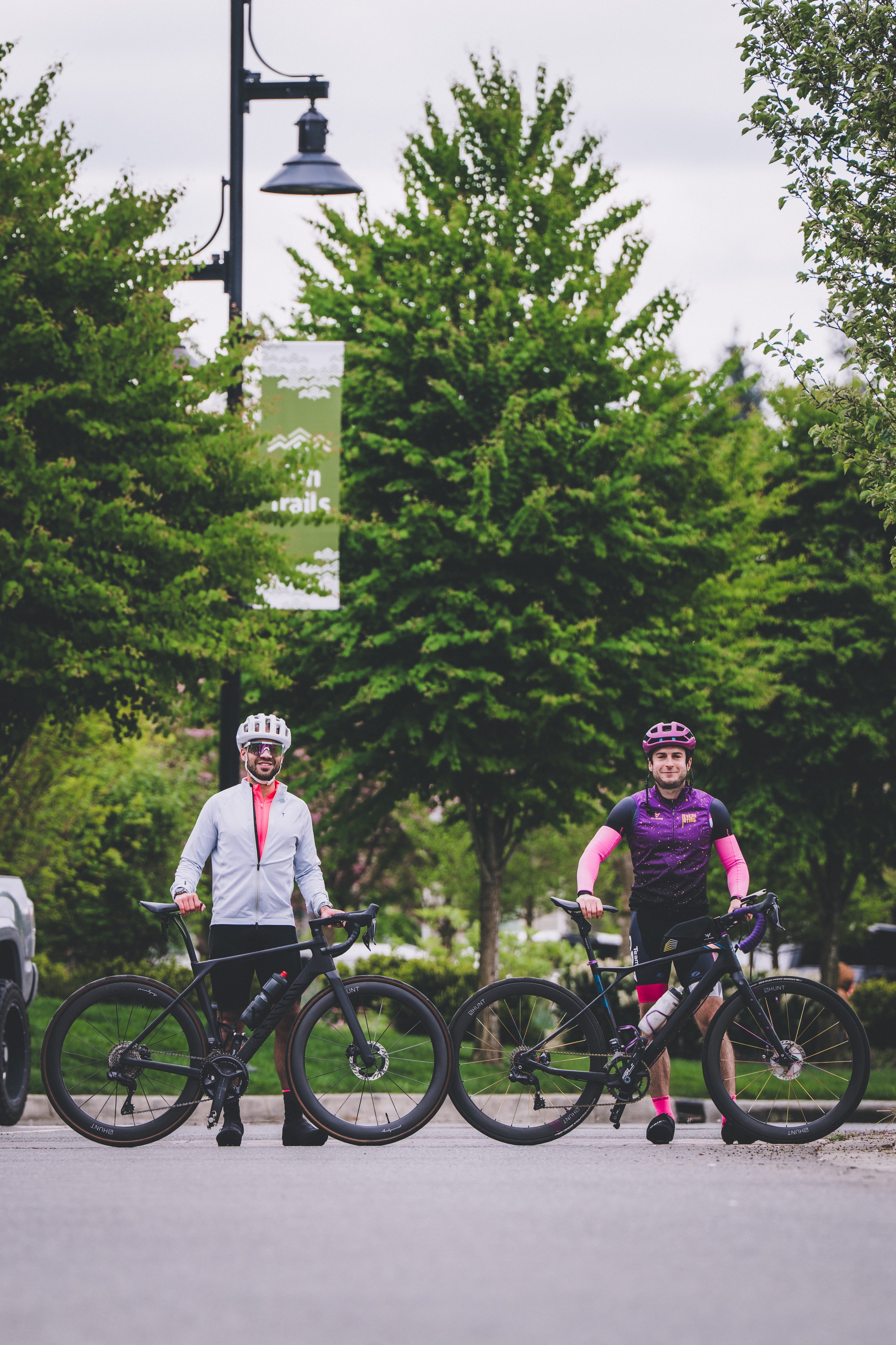 Two cyclists wearing helmets and cycling gear stand with their bikes on a paved path, surrounded by lush green trees
