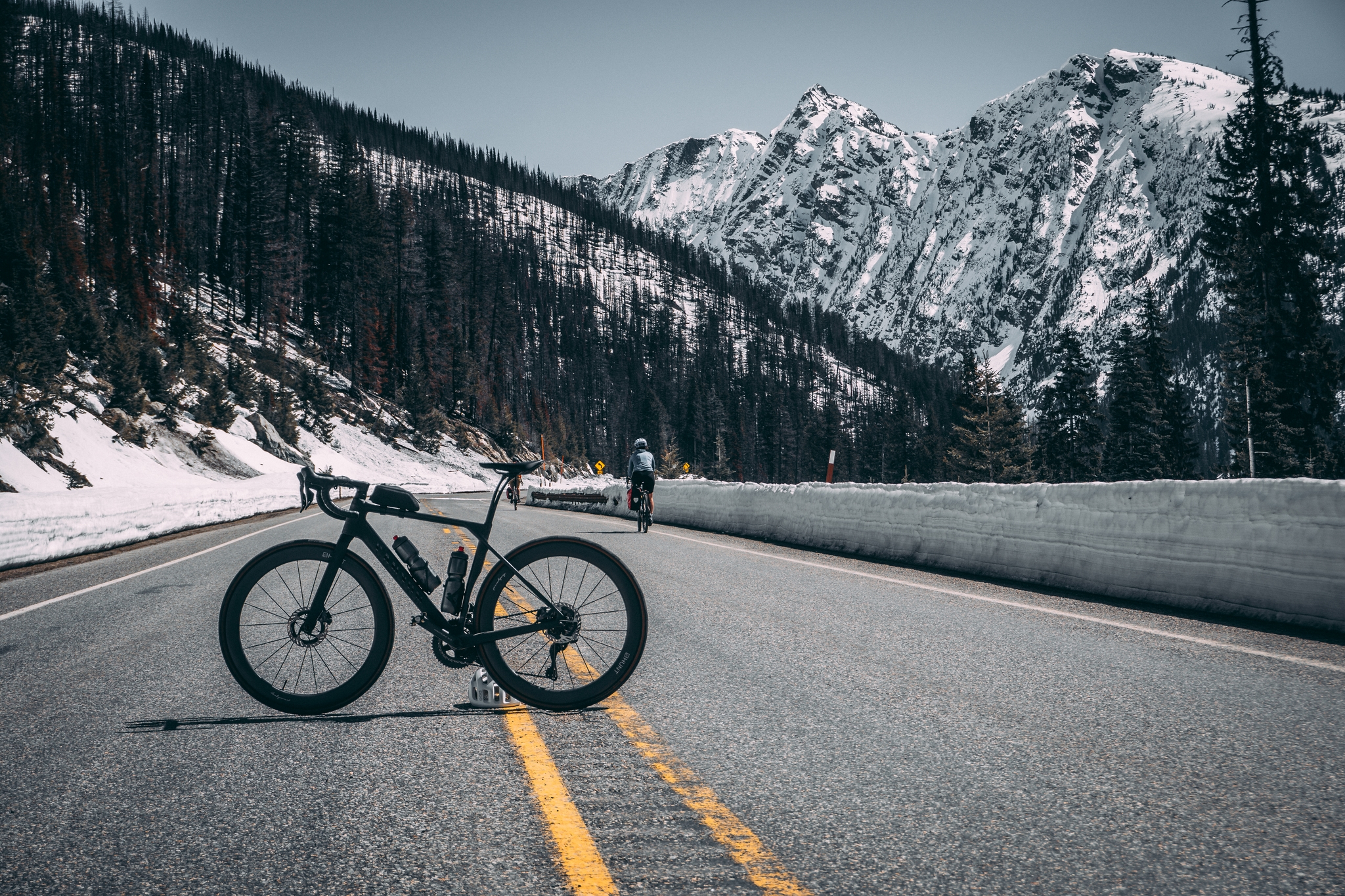 A canyon endurace is parked on a mountain road with snow-covered peaks and pine trees in the background
