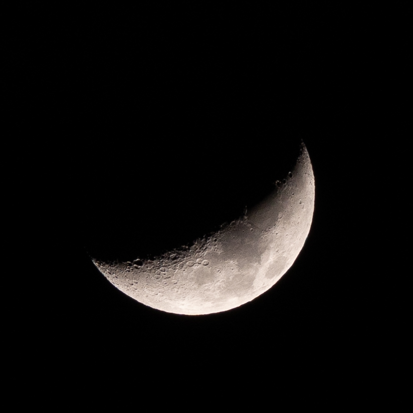 A bright crescent moon hangs against a black night sky, its illuminated curve revealing subtle craters and surface texture