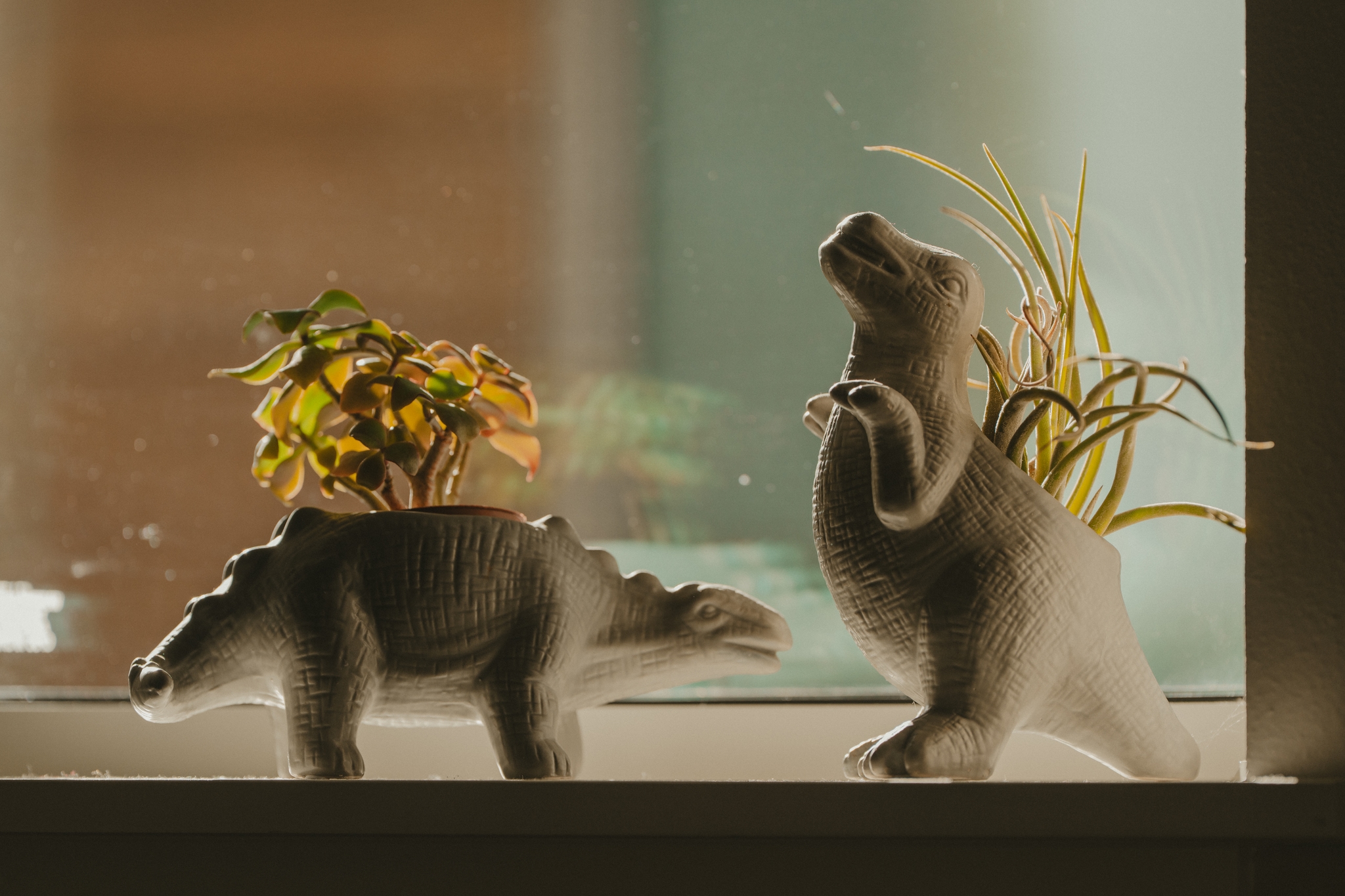 Two dinosaur-shaped planters with small plants on their backs, placed on a windowsill