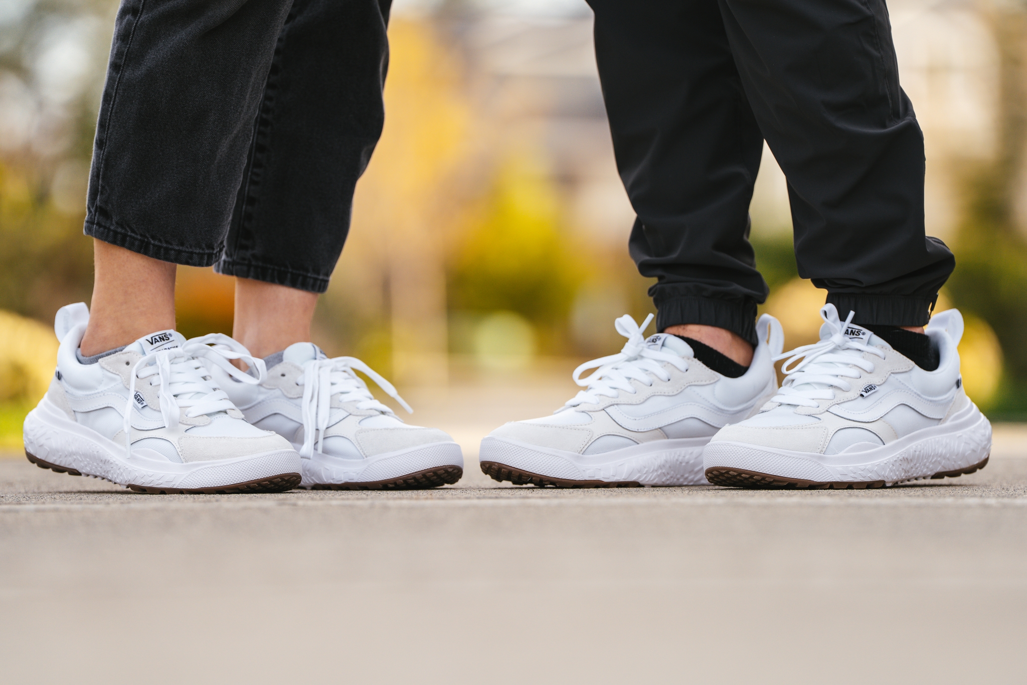 Two people wearing white sneakers stand facing each other on a paved surface, with blurred greenery in the background