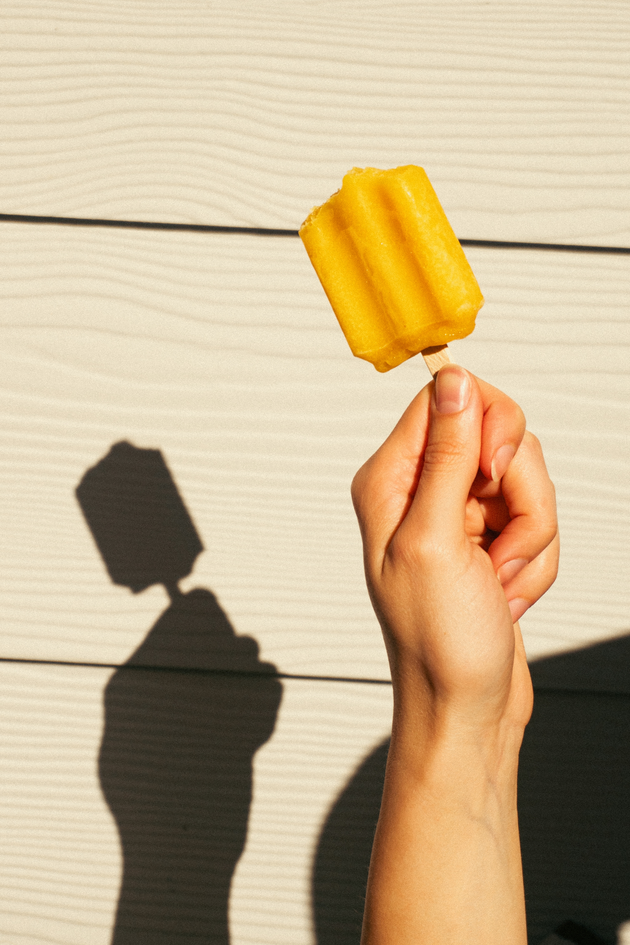 A hand holding a yellow popsicle against a light-colored wall, casting a shadow