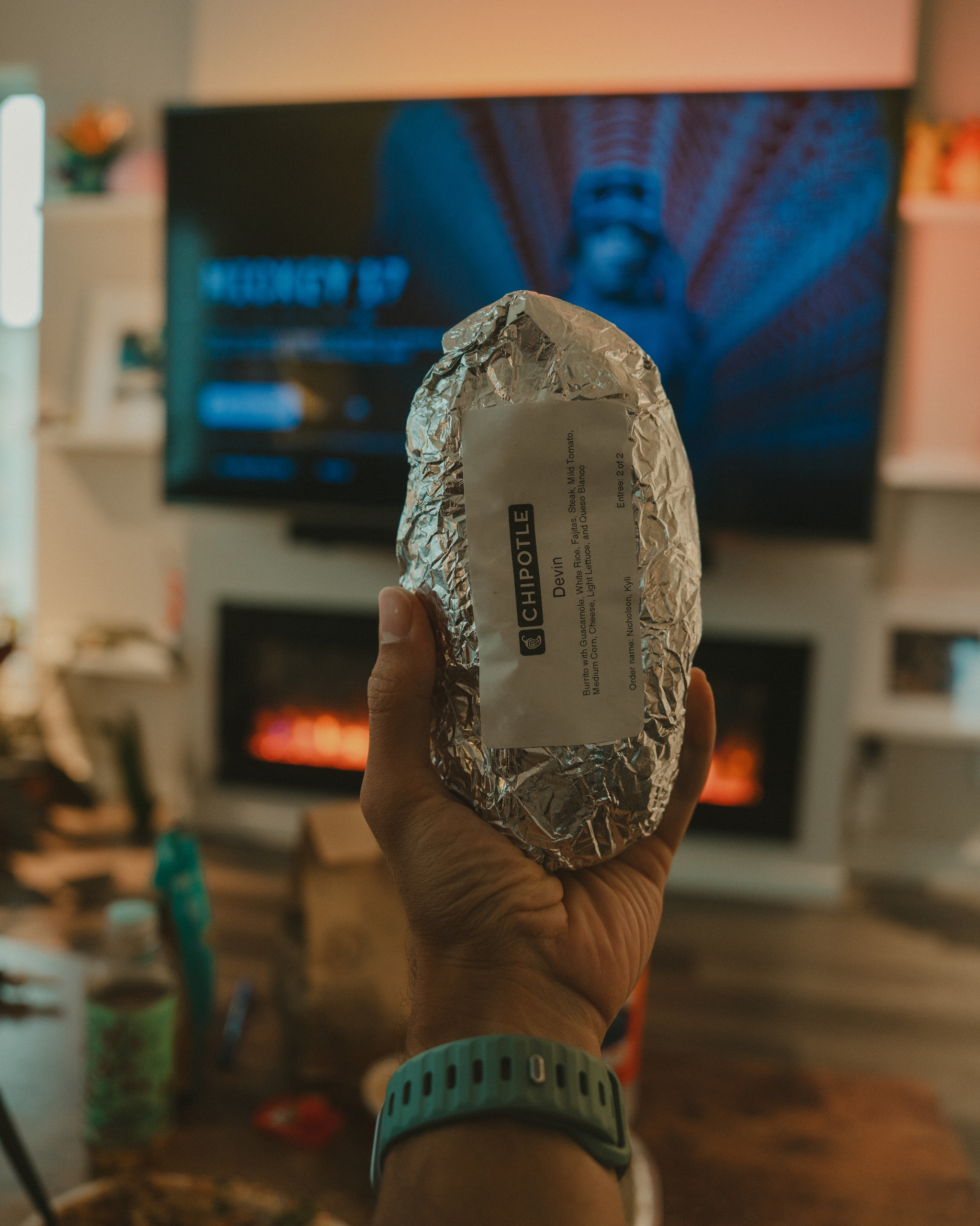A hand holding a foil-wrapped burrito in a living room with a TV and fireplace in the background