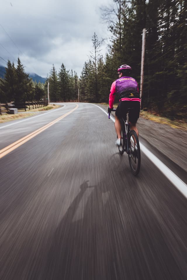 A cyclist wearing a pink jacket and helmet rides along a winding road surrounded by trees, with a cloudy sky overhead
