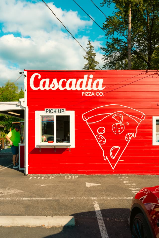 A red building with Cascadia Pizza Co written on it, featuring a large pizza slice illustration and a pick-up window