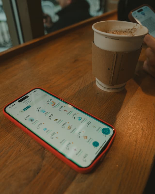 A smartphone with a red case displaying a messaging app rests on a wooden table beside a paper coffee cup. A hand is holding another phone in the background