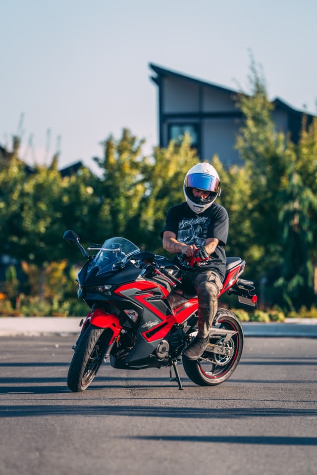 A person wearing a helmet sits on a red motorcycle, parked on a paved surface, with trees and a building in the background