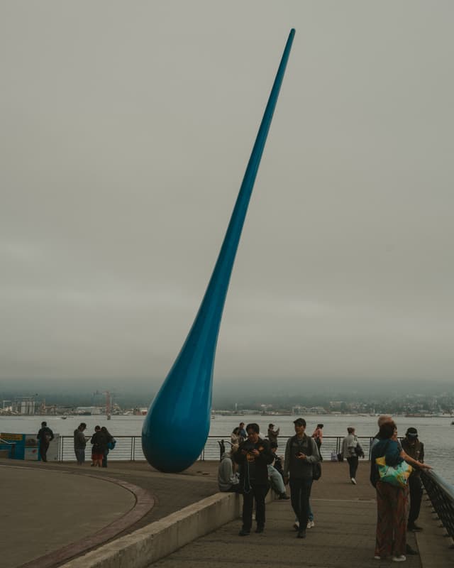 Tall blue raindrop sculpture by waterfront with people walking around