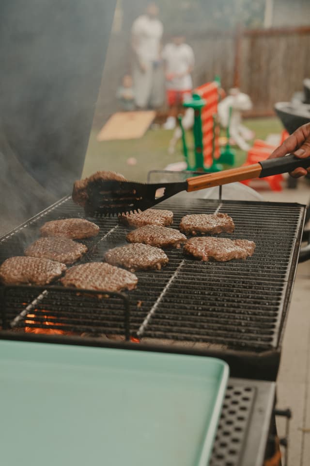 Grilling burgers on an outdoor barbecue, with smoke rising and people in the background
